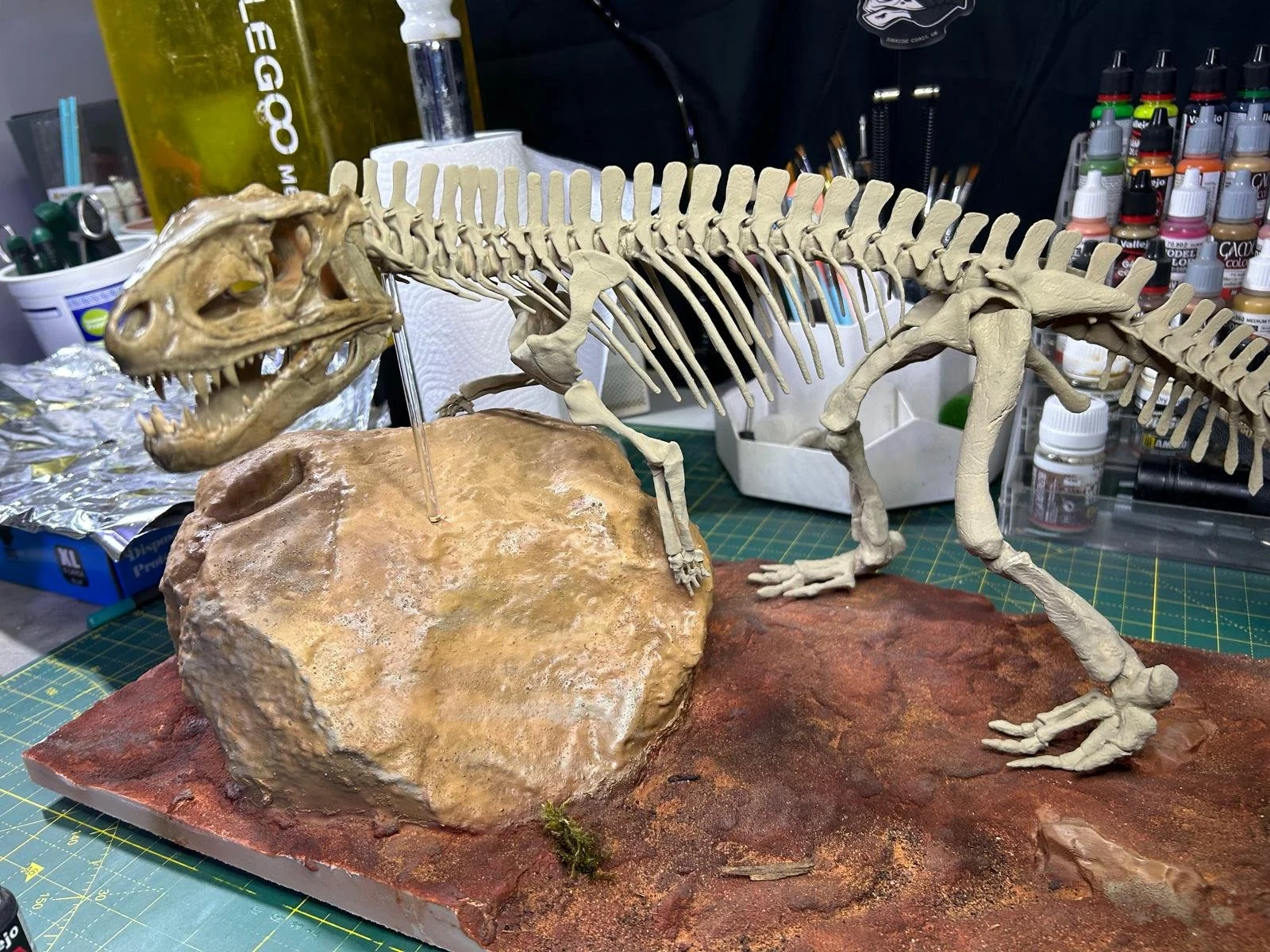A detailed postosuchus skeleton model, is displayed on a workspace. The skeleton is positioned on a rocky base with tools and paints in the background, indicating a model-building or paleontology setting.