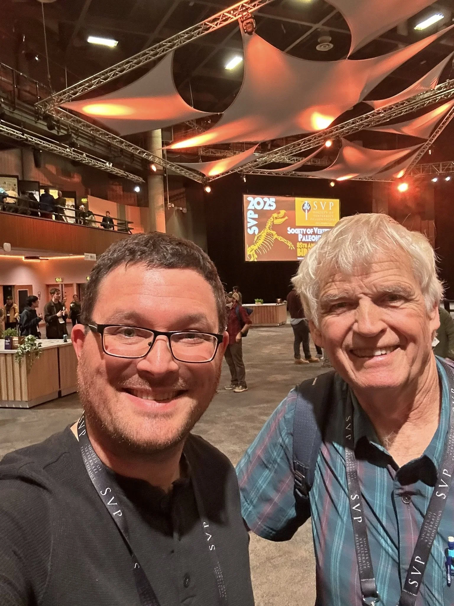 Two men smiling for a selfie in an event hall with a high ceiling, orange lighting, and a large screen displaying 'SVP 2025 Society of Vertebrate Paleontology.