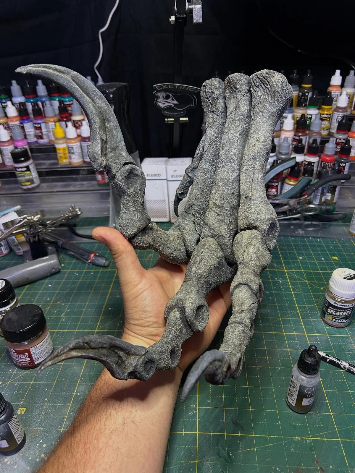 A person holding a large, detailed, sculpted model of a utahraptor foot, with hobby paints and art supplies in the background on a craft table.