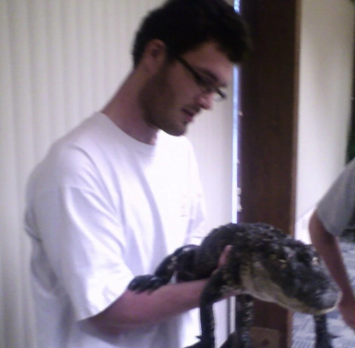 A young man with dark hair, glasses, wearing a white shirt, holding a large, dark-colored alligator.