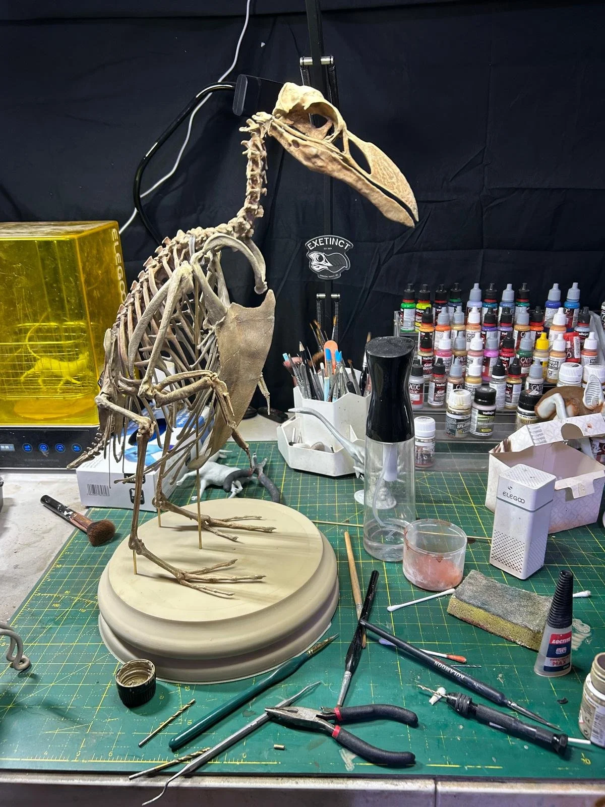 A workspace of a taxidermist with a partially assembled great auk skeleton, various paint bottles, tools, and art supplies on a green cutting mat.