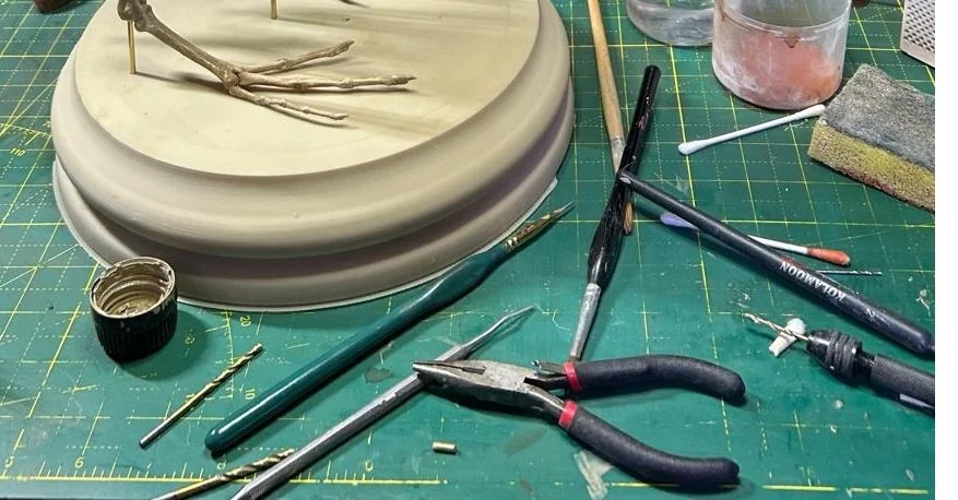 Assorted crafting tools and supplies on a green cutting mat, including pliers, paintbrushes, a sponge, small containers, and a sculpture base with a small branch or twig.