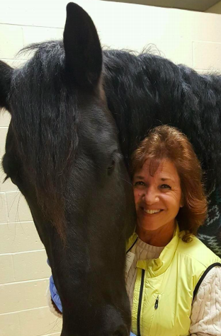 A woman smiling next to a large black horse in an indoor stable.