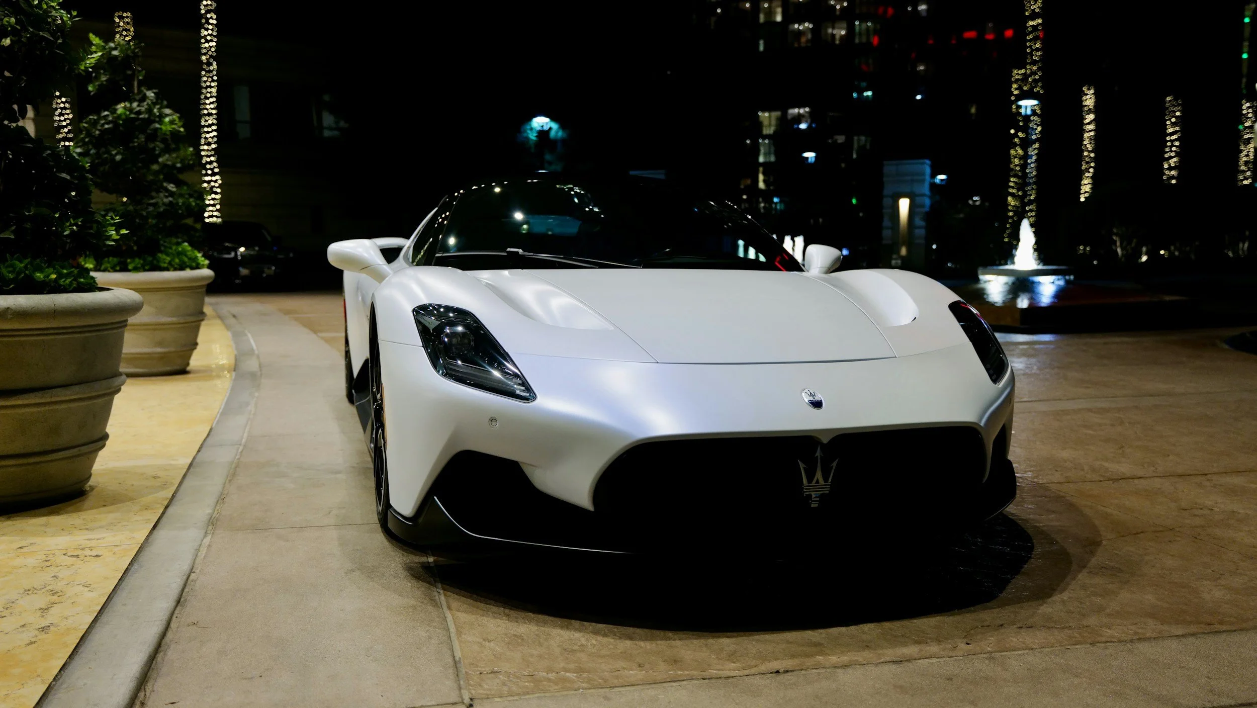 White sports car parked on a paved area at night, with potted plants on the left, illuminated buildings and a fountain in the background.