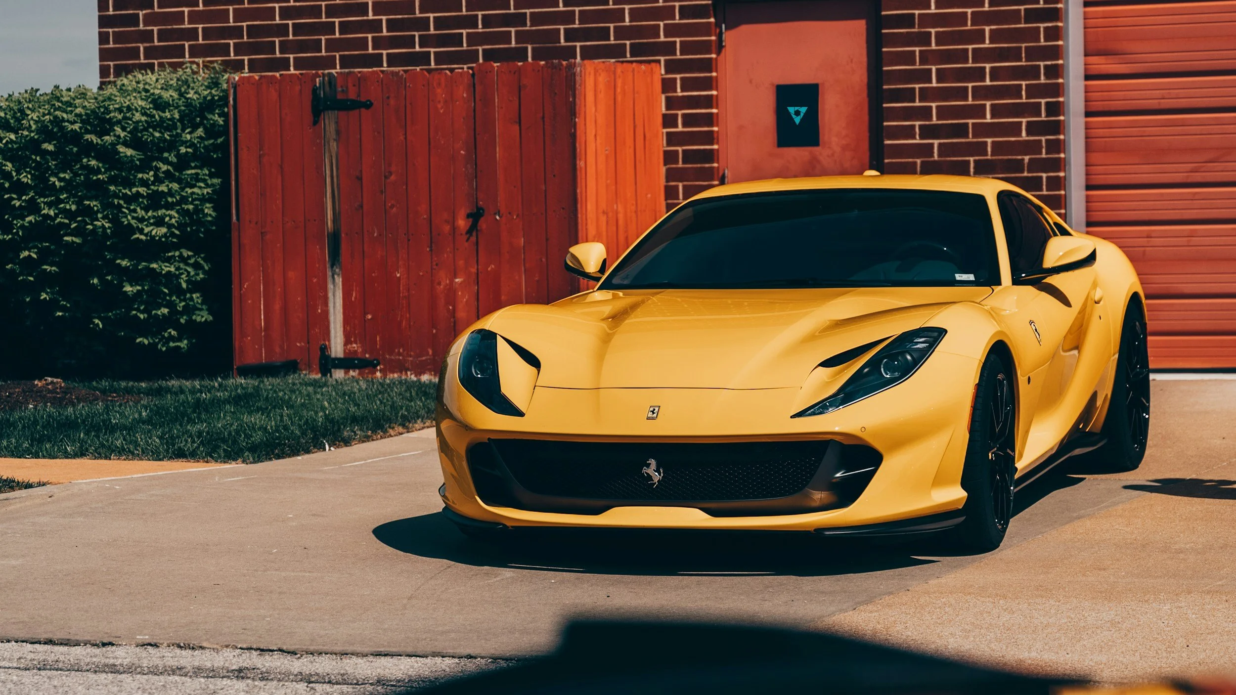 A yellow Ferrari sports car parked on a driveway near a red brick building with a wooden fence and green bushes in the background.
