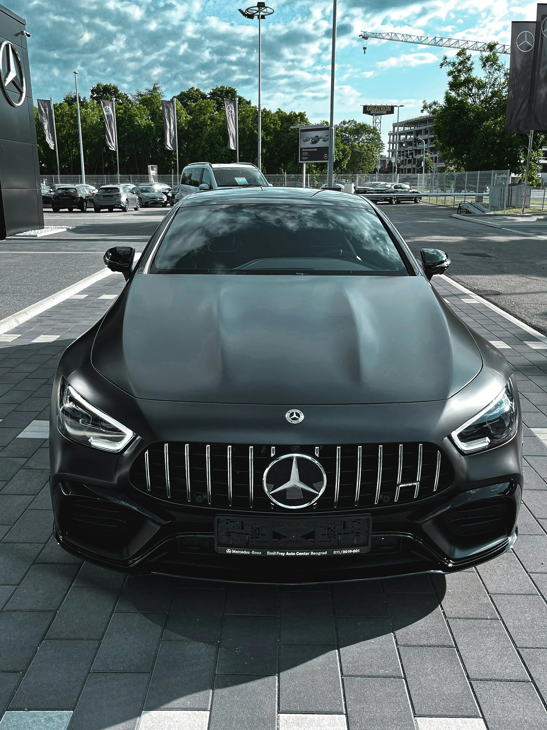 Black Mercedes-Benz car parked in a dealership lot with other cars in the background. The front view shows the Mercedes logo on the grille and reflective windshield.