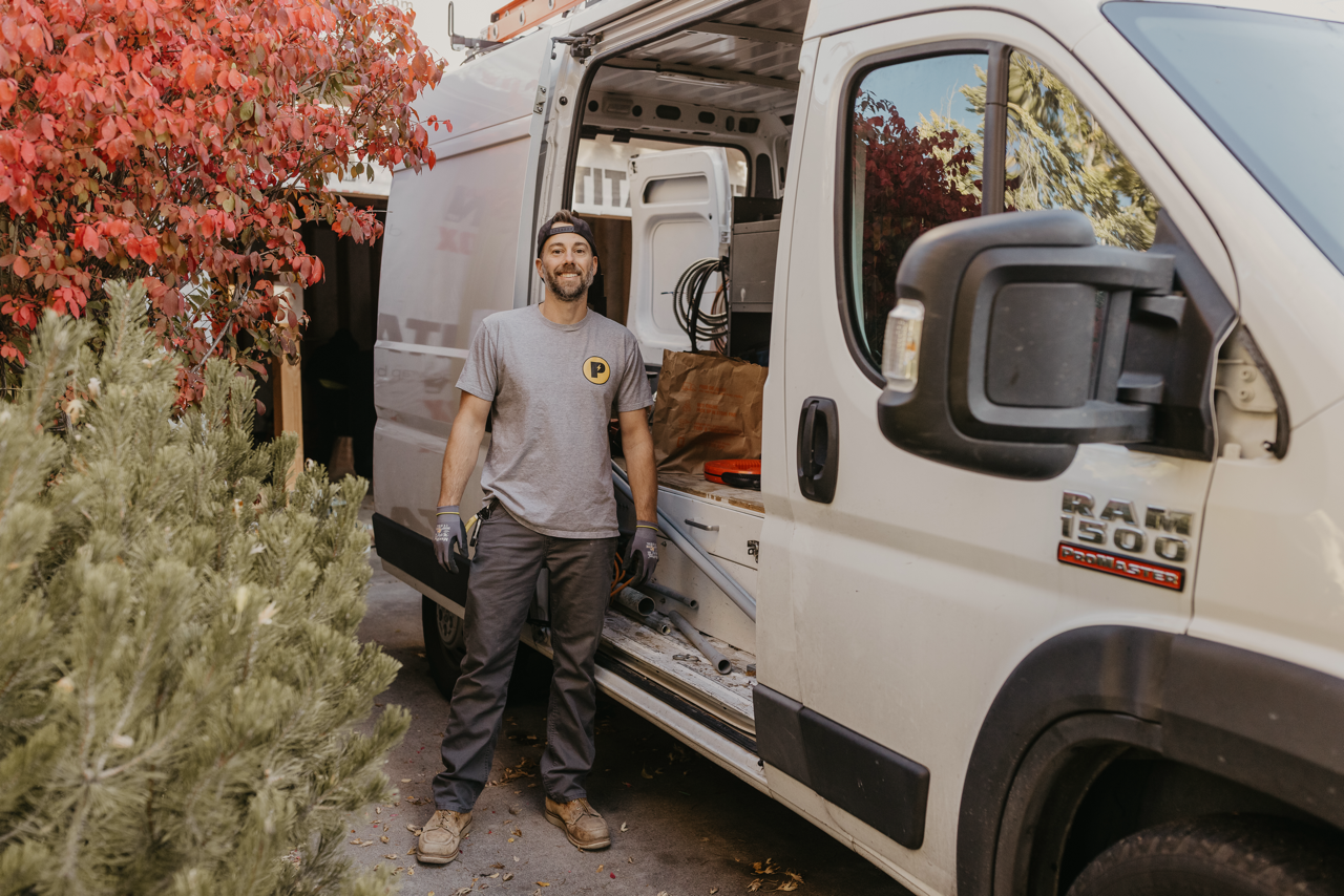A man stands outside a work van, smiling, with tools and equipment inside. He is wearing a gray t-shirt, black pants, work gloves, and a cap. The van is parked beside trees with red and green leaves.