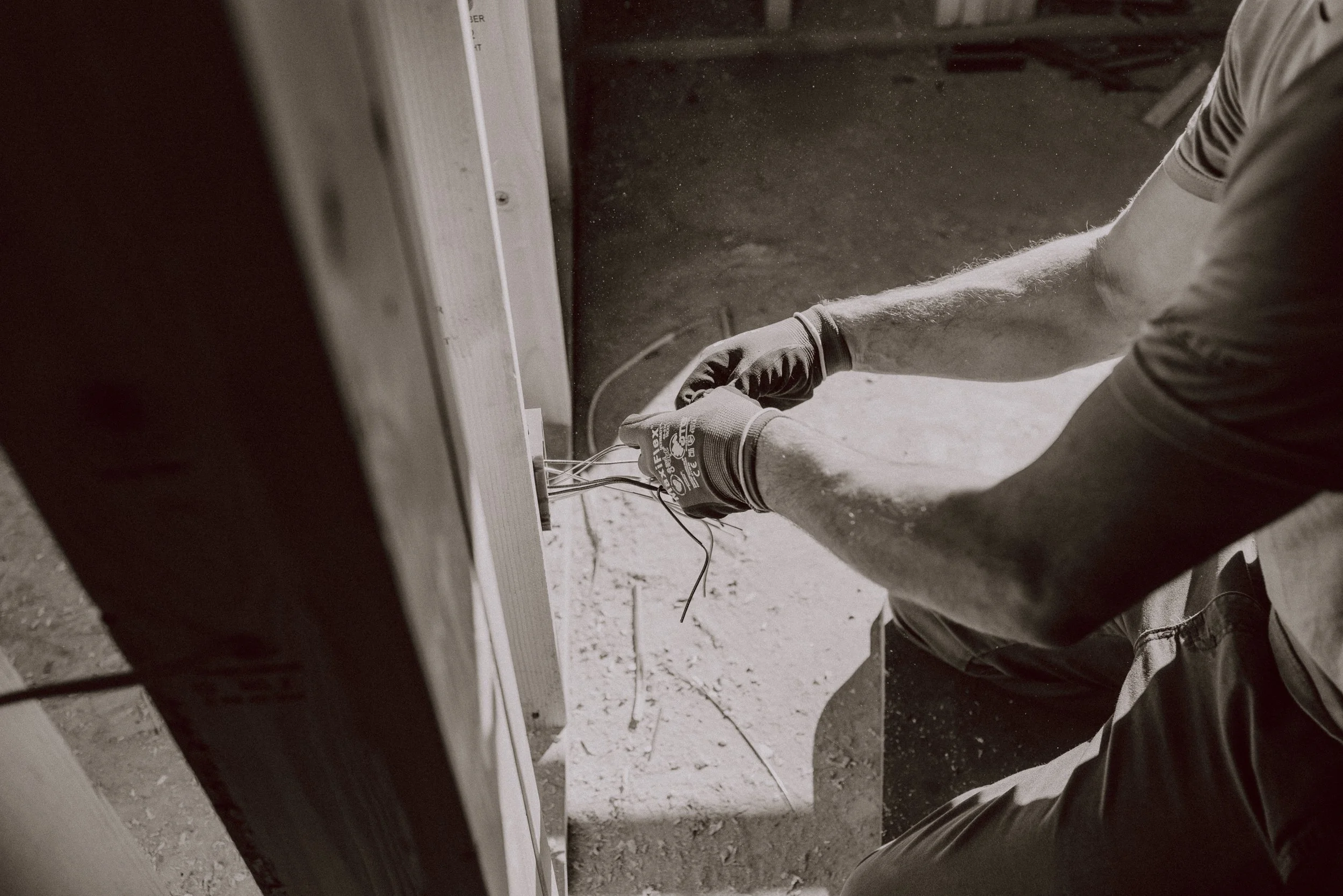 A person working on electrical wiring at a construction site, wearing gloves and handling multiple wires. The image is in black and white.