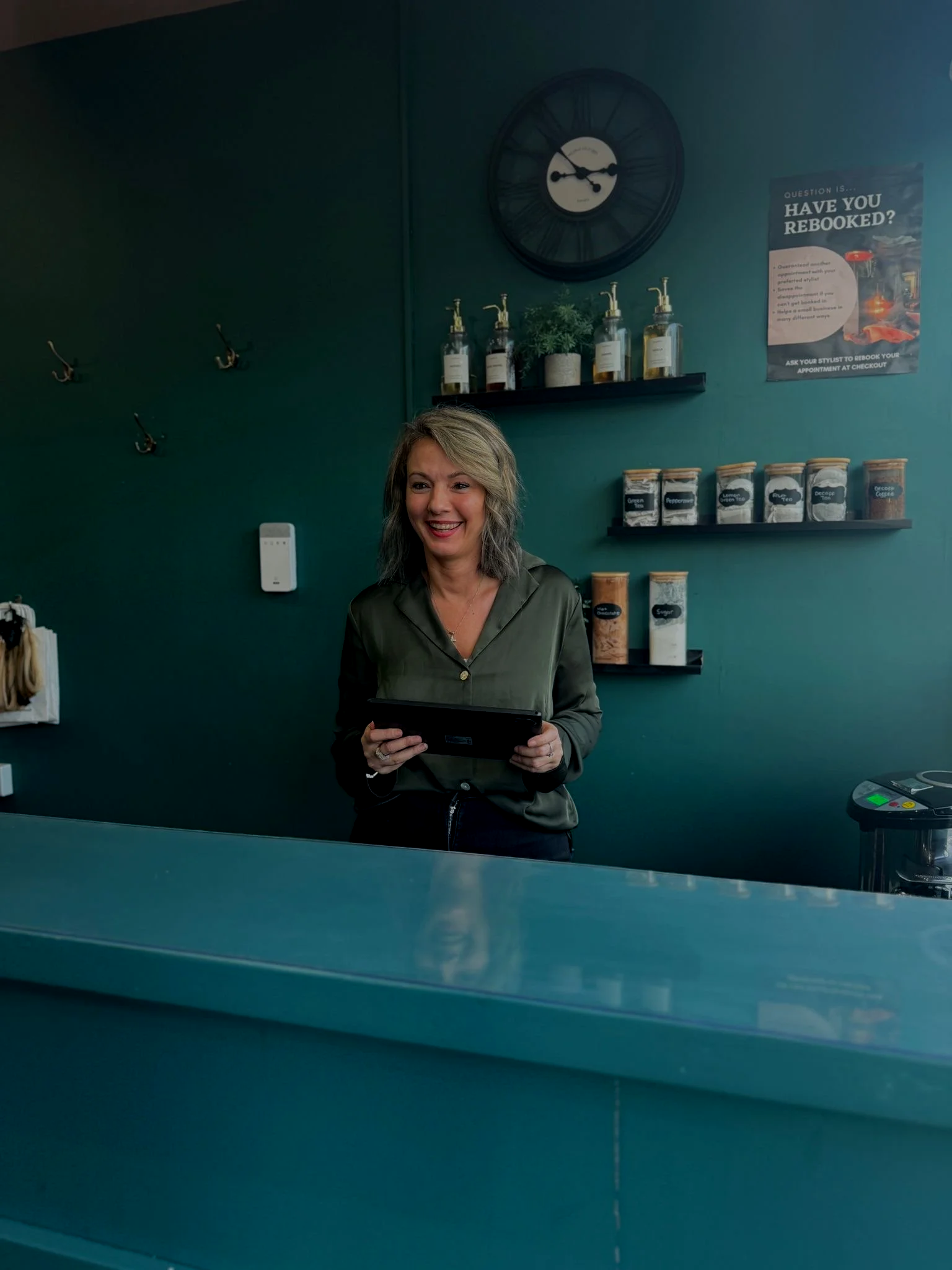 A woman standing behind a counter in a retail or cafe setting, smiling and holding a tablet. The background has teal walls, black shelves with spice jars, and a clock on the wall.