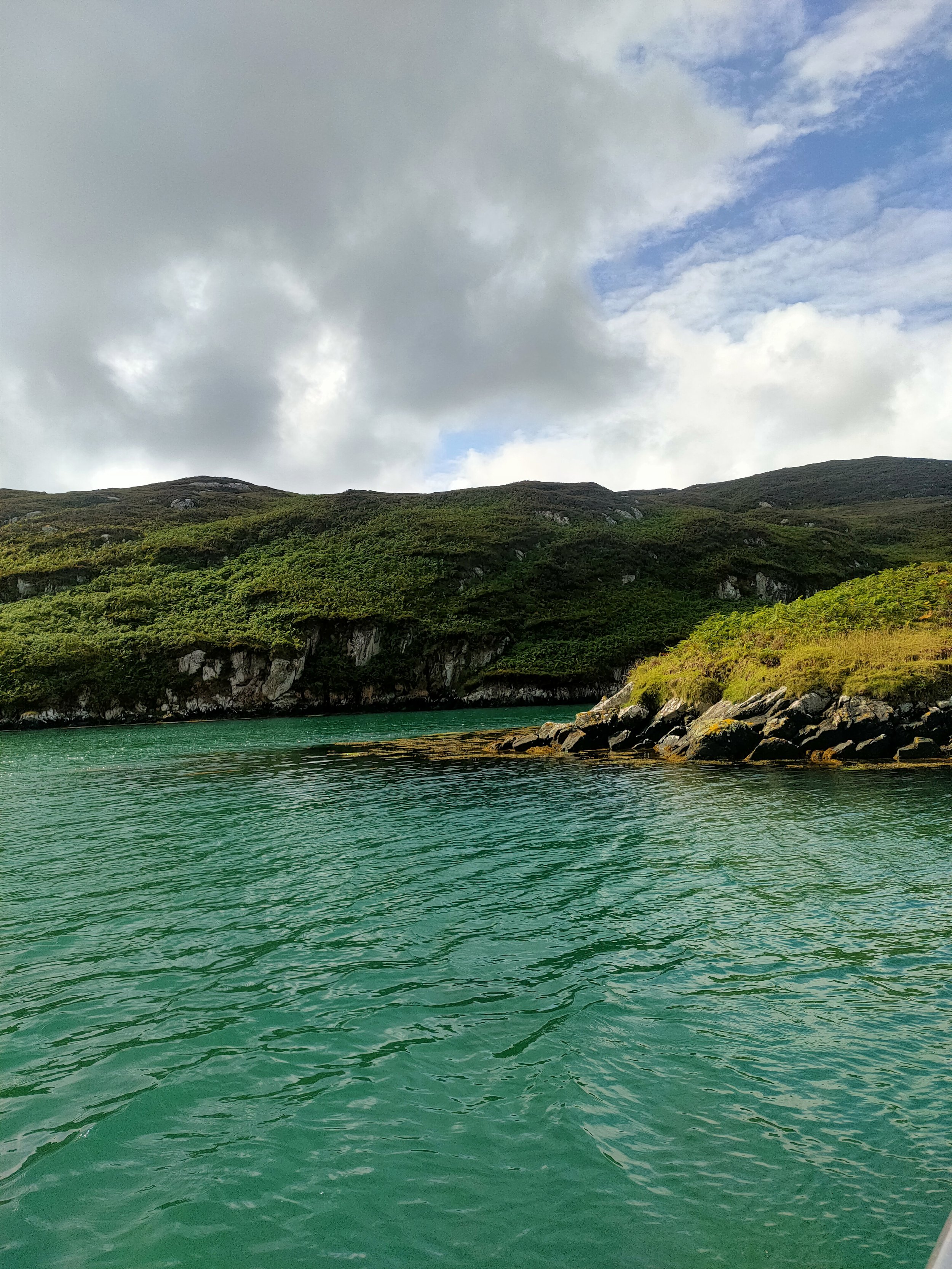 A serene body of turquoise water surrounded by green hills with rocky cliffs, under a partly cloudy sky.