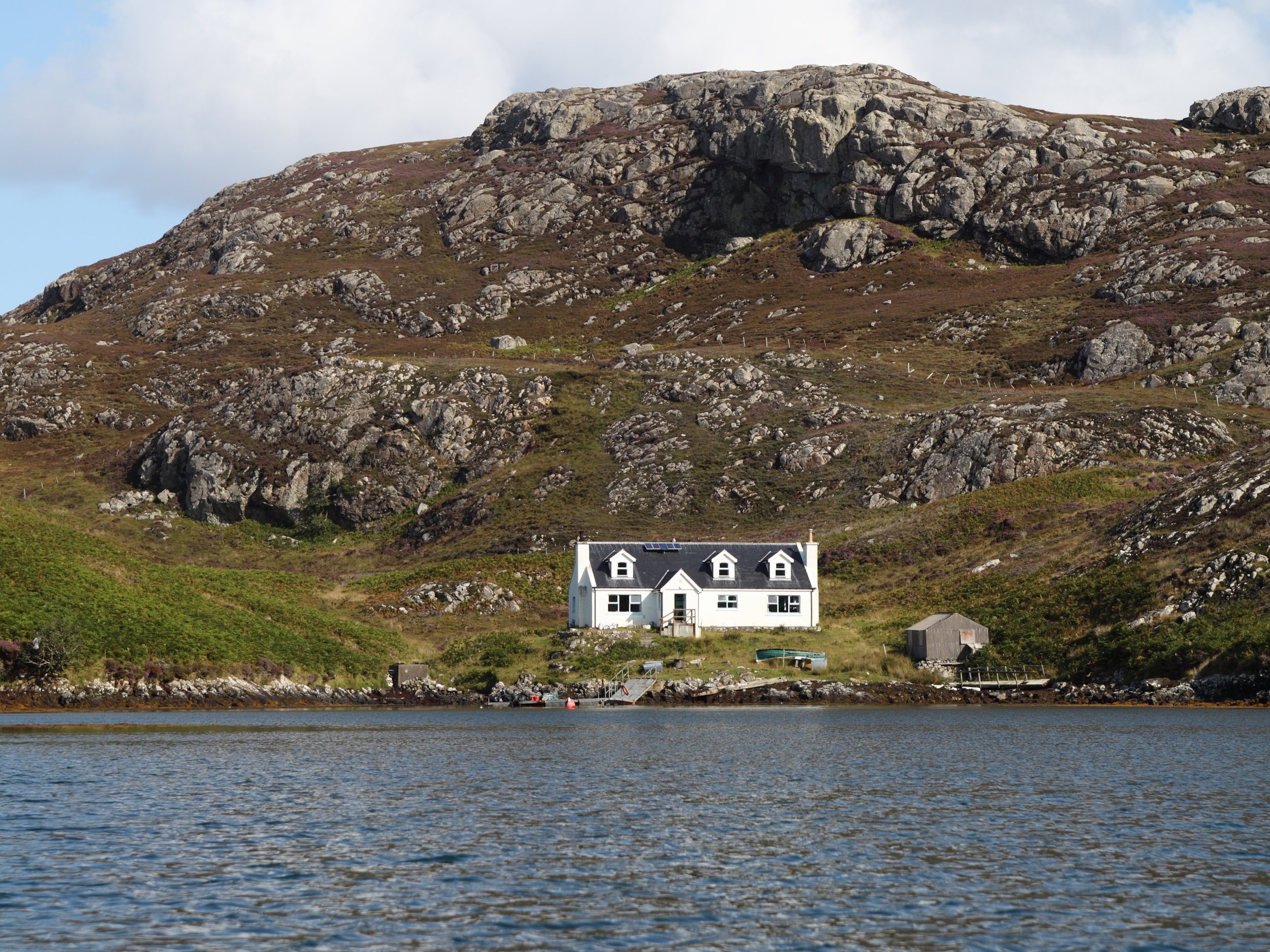 House on the shoreline with a rocky hillside in the background and water in the foreground.
