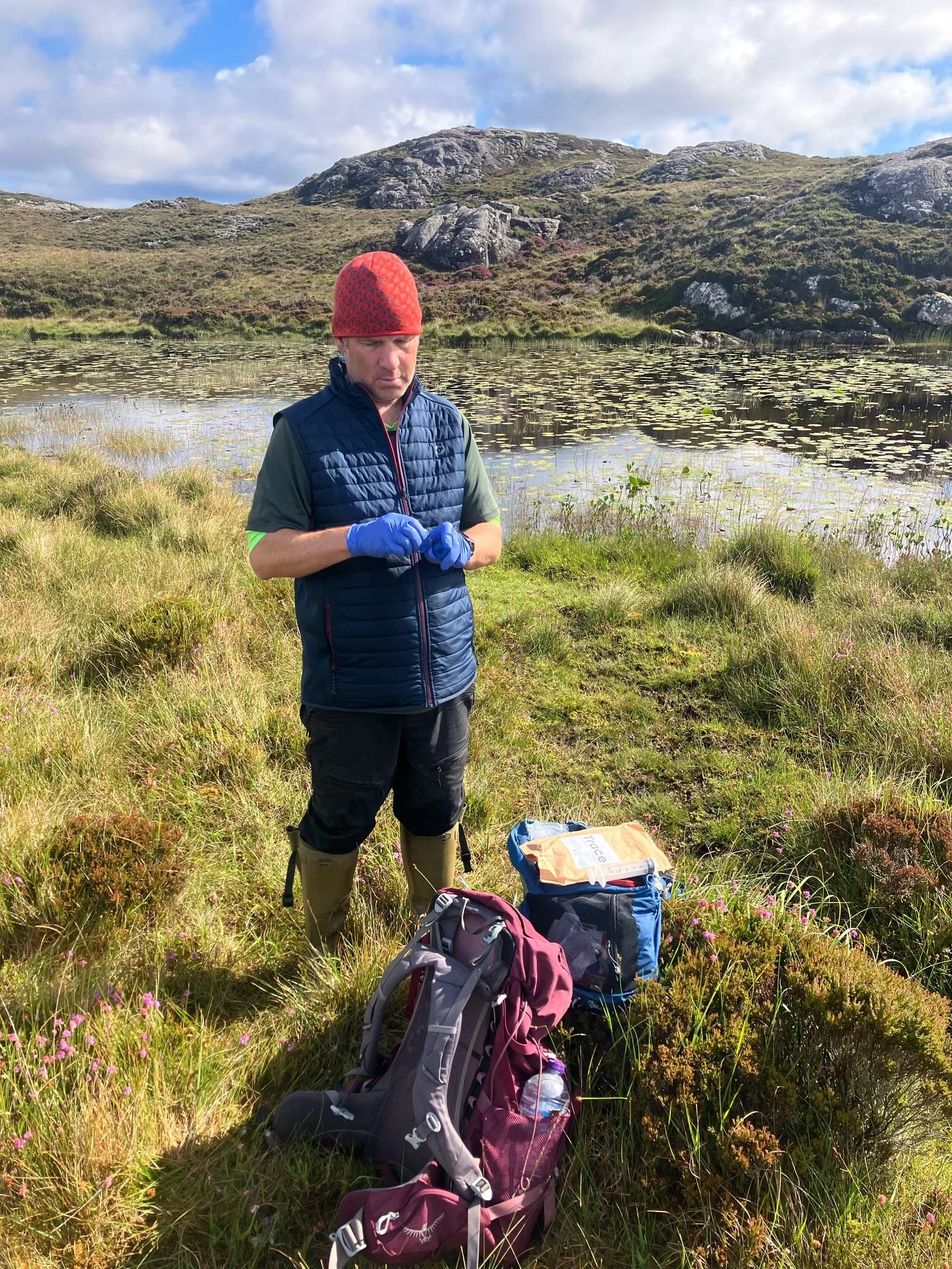 Man in outdoor gear standing on a grassy slope, holding a walking stick, with rolling hills in the background.