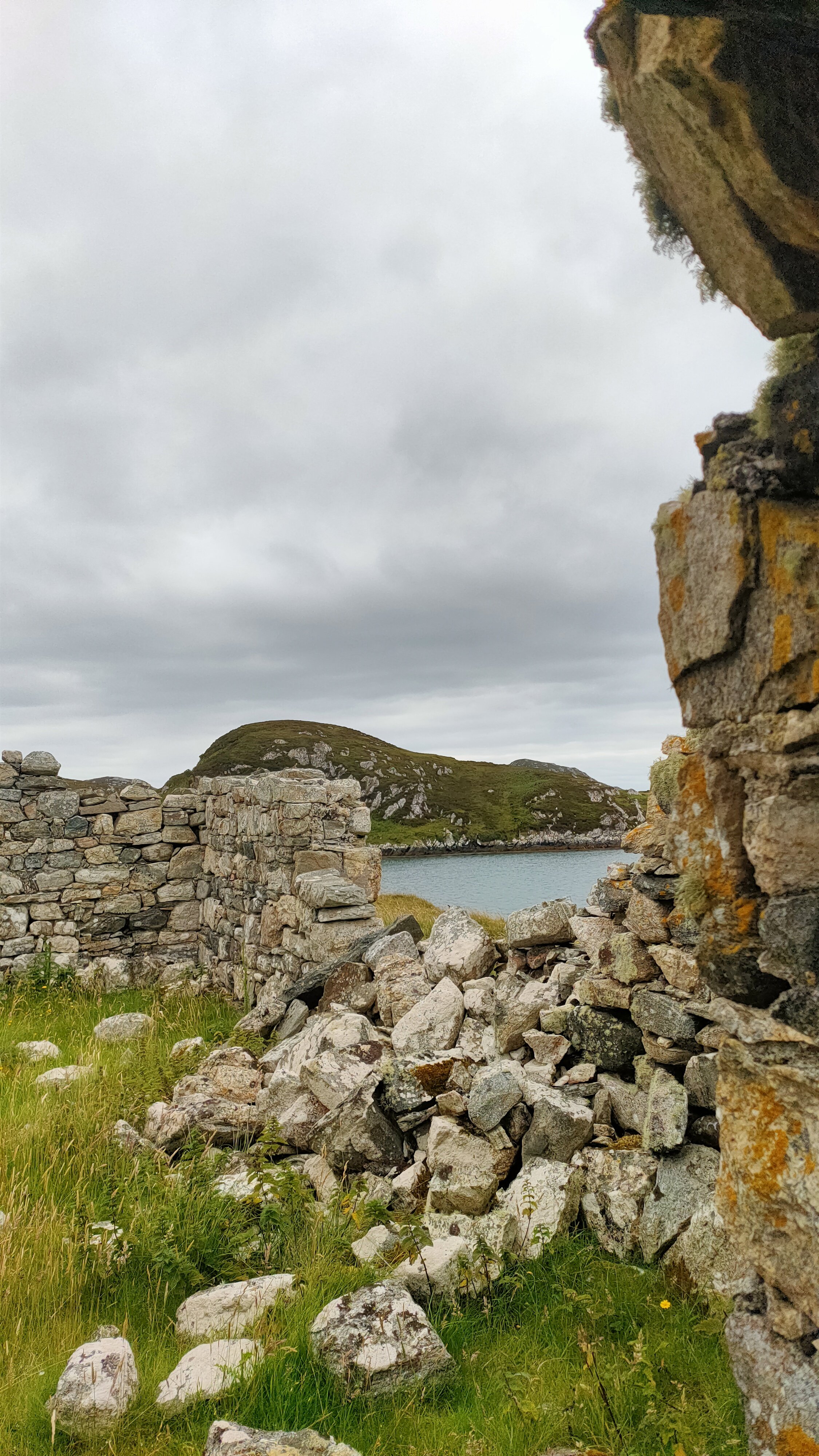 Ruins of stone walls in a grassy landscape with a hill and water body in the background, under a cloudy sky.