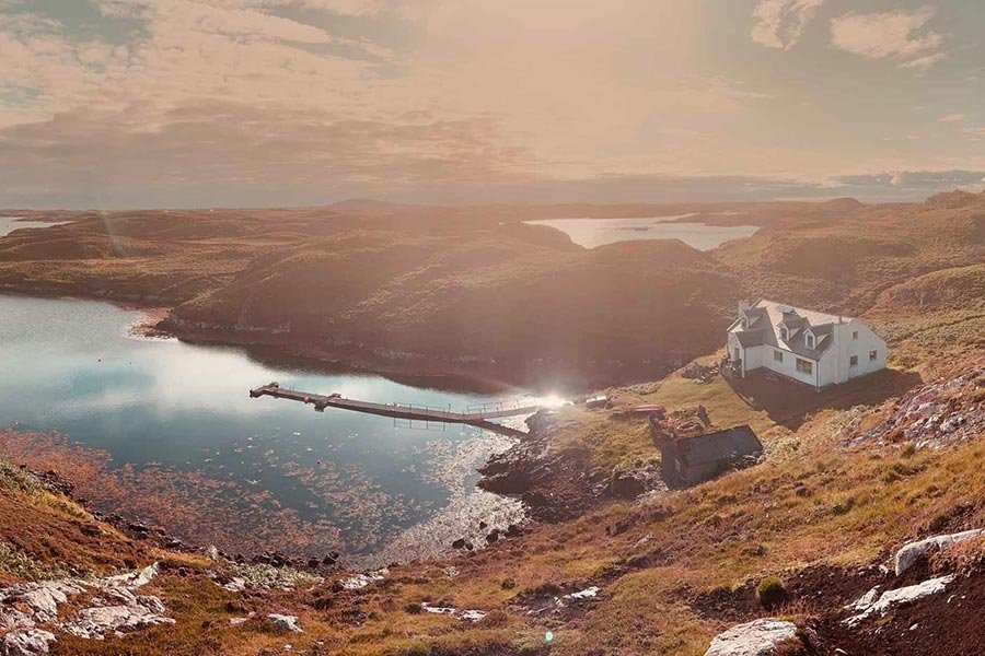 Scenic view of a lake surrounded by hills, with a white house and smaller building on the hillside, dock extending into the lake, and partly cloudy sky at sunset.