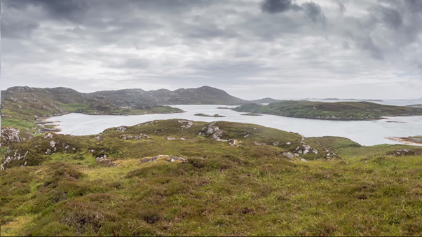 Scenic view of a landscape with rolling green hills, lakes, and distant mountains under a cloudy sky.