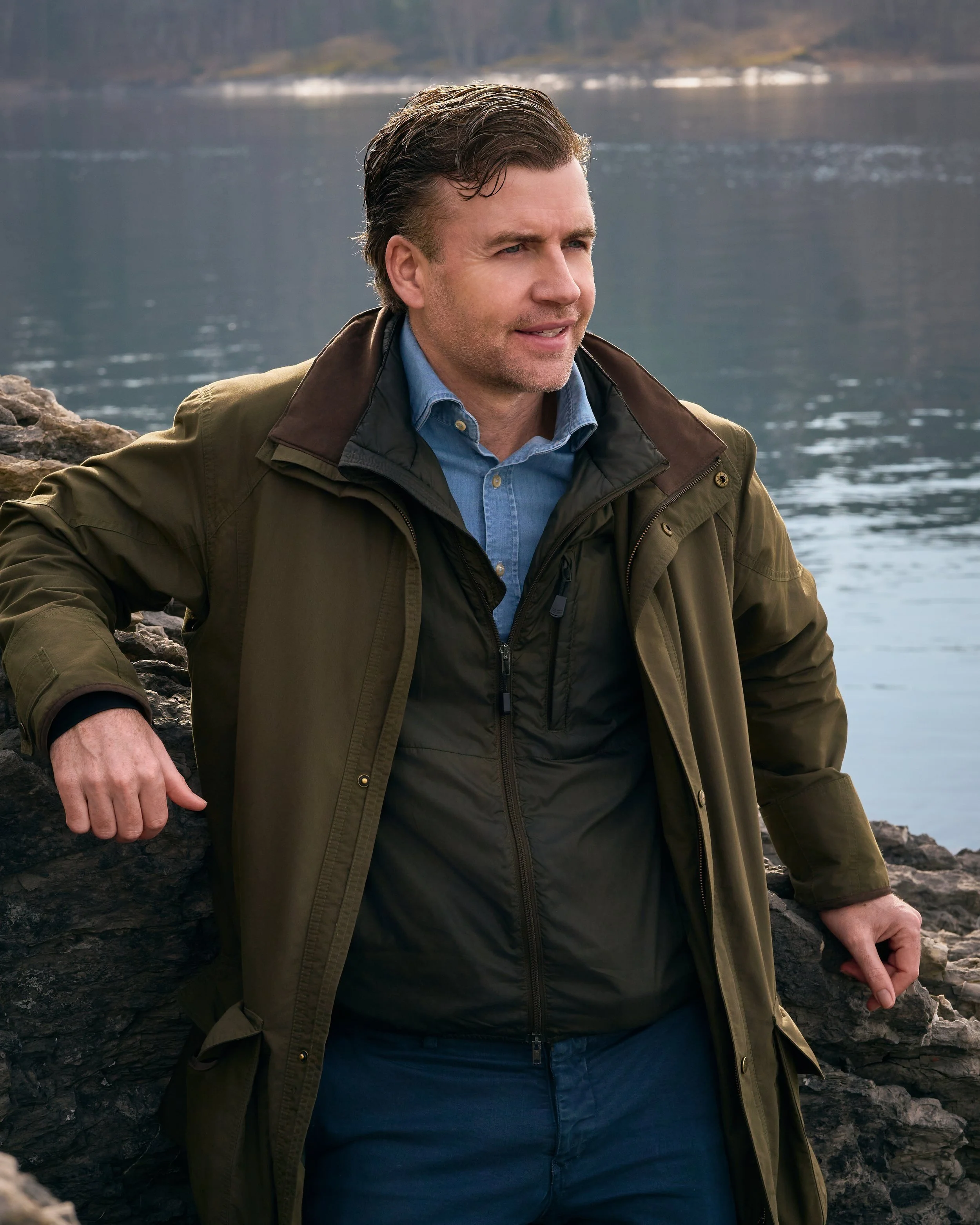 Andrew Marshall standing outdoors near a body of water, with rocky terrain in the background, wearing a green and black jacket over a blue shirt.
