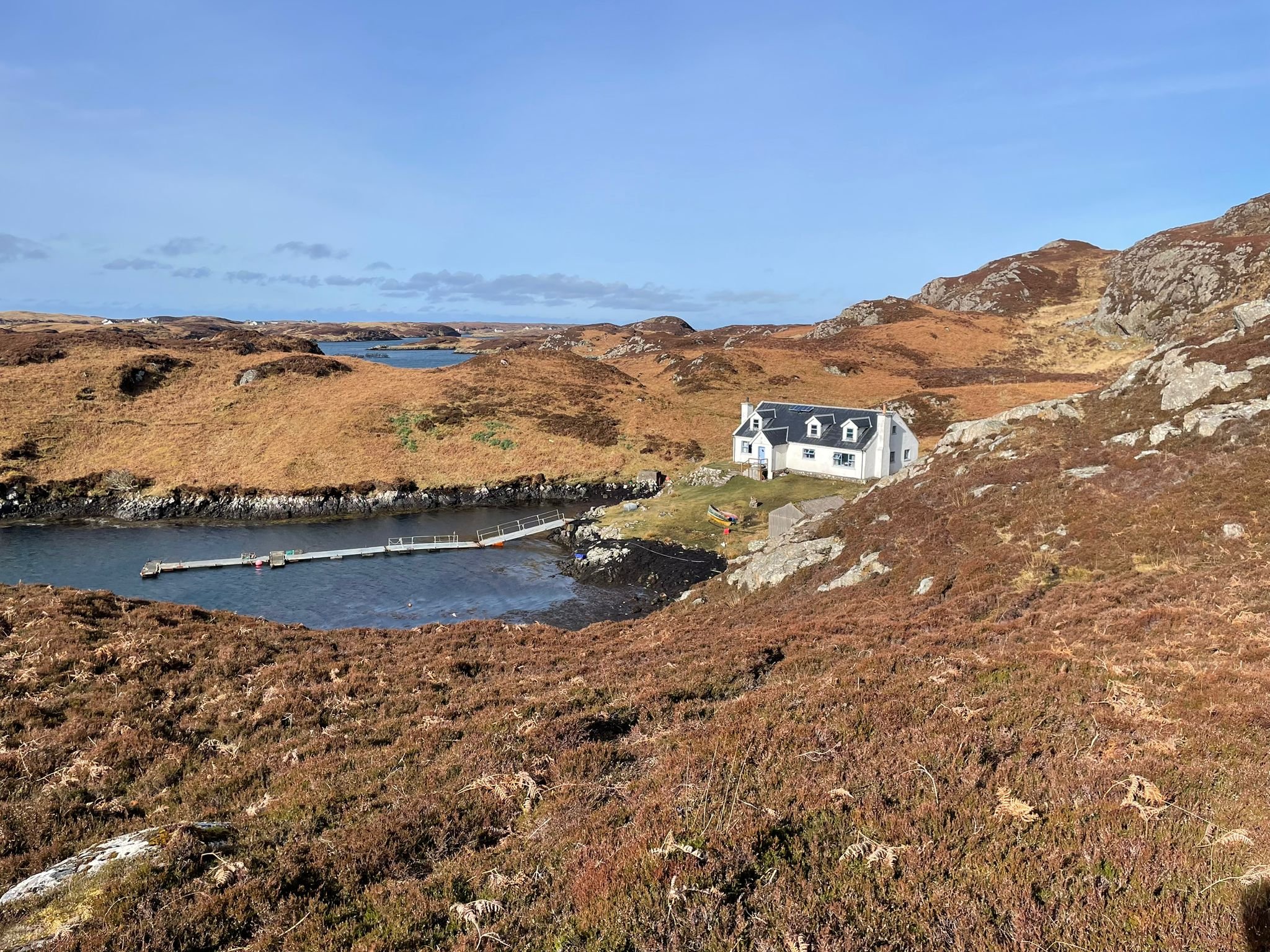 Scenic landscape with a white house near a small harbor, surrounded by rolling brown hills and a bright blue sky.
