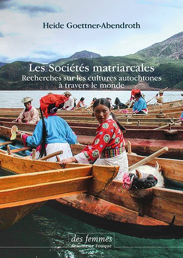 Femmes autochtones au bord d'un lac, assises dans des bateaux en bois, portant des vêtements traditionnels colorés, entourées de montagnes.
