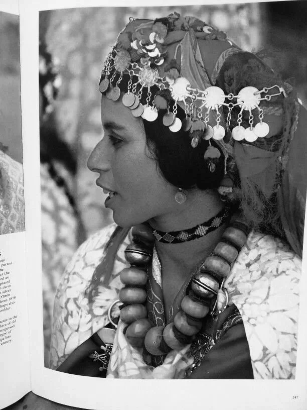 Portrait d'une femme portant des bijoux traditionnels, un chapeau orné de pièces métalliques suspendues, des colliers volumineux en bois et accessoires, vêtements traditionnels et décoratifs.