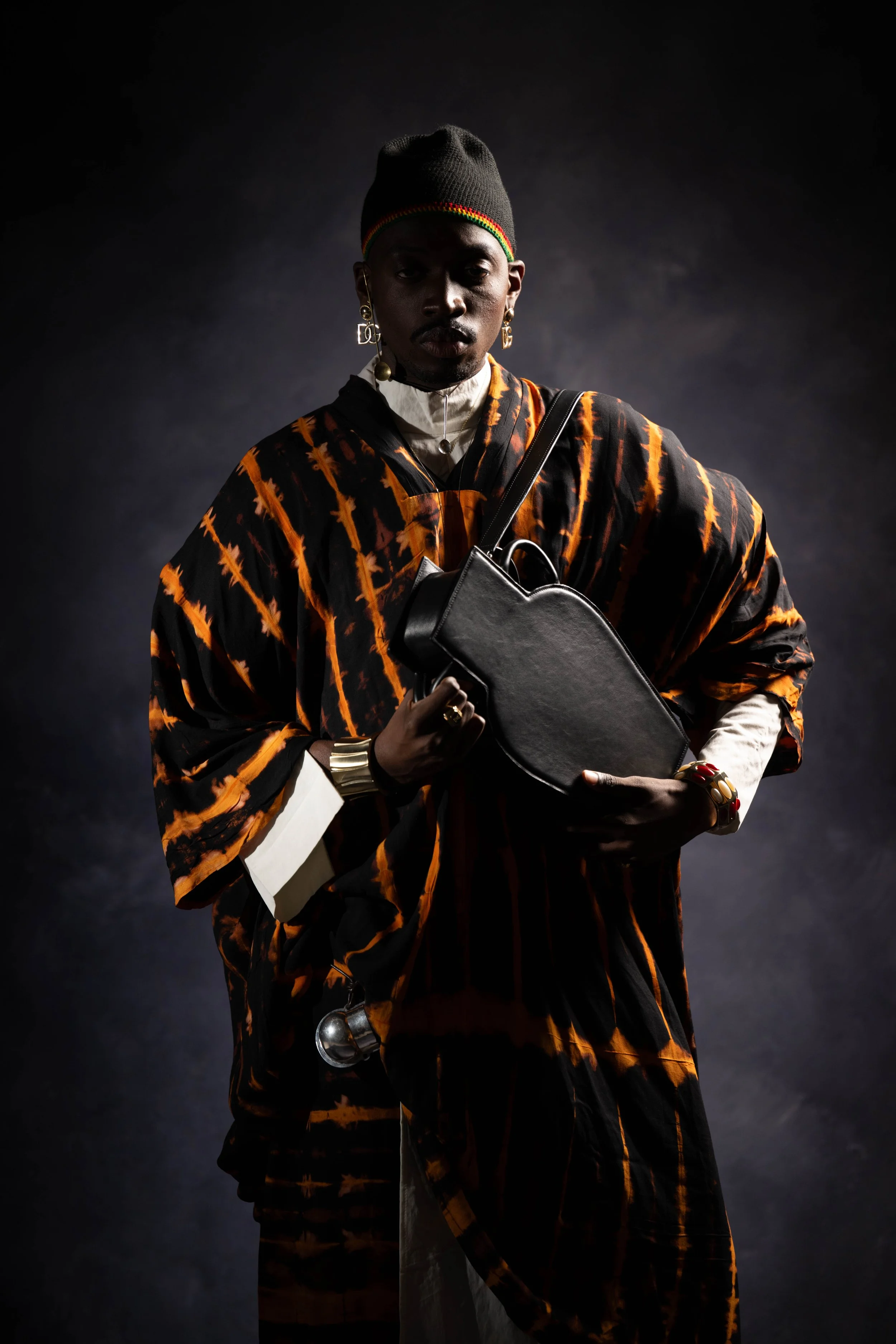 Personne portant un vêtement à motif orange et noir, un bonnet noir, tenant un sac à main noir, décorative et portant des bijoux, devant un fond sombre.