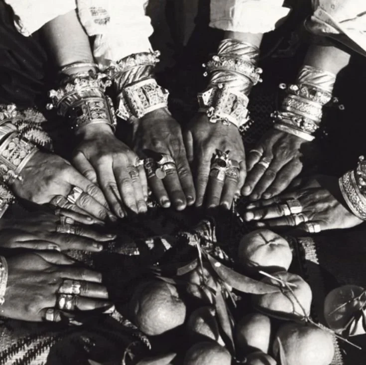 Groupe de plusieurs mains portant de nombreux bijoux, entourant un objet au centre, avec des fruits autour.