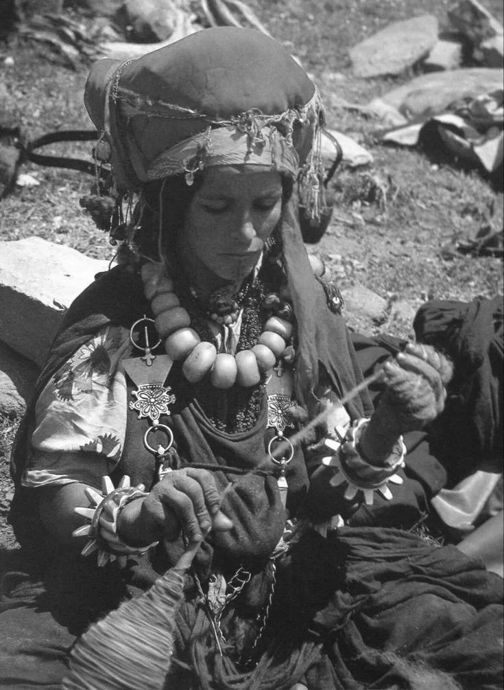Une femme portant des vêtements traditionnels, des bijoux et un headgear, tenant un outil, dans un environnement extérieur avec des rochers.
