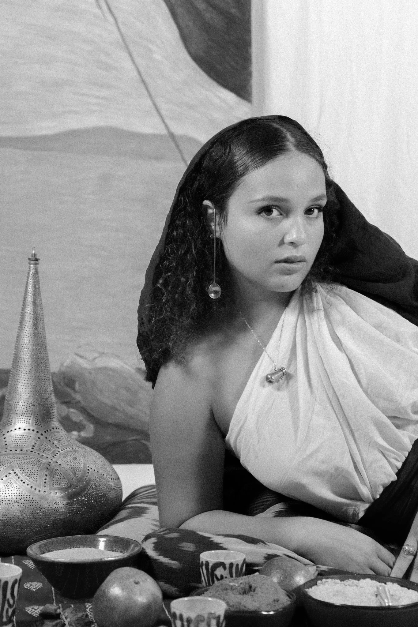 Une femme aux cheveux bouclés portant une robe blanche, avec des bijoux, assise à une table avec des objets décoratifs, des fruits et des bols, dans une pièce avec une œuvre d'art en arrière-plan.