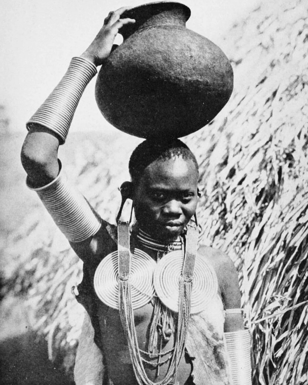 Femme africaine portant de multiples bijoux traditionnels et un grand vase sur la tête, dans un champ de cultures.
