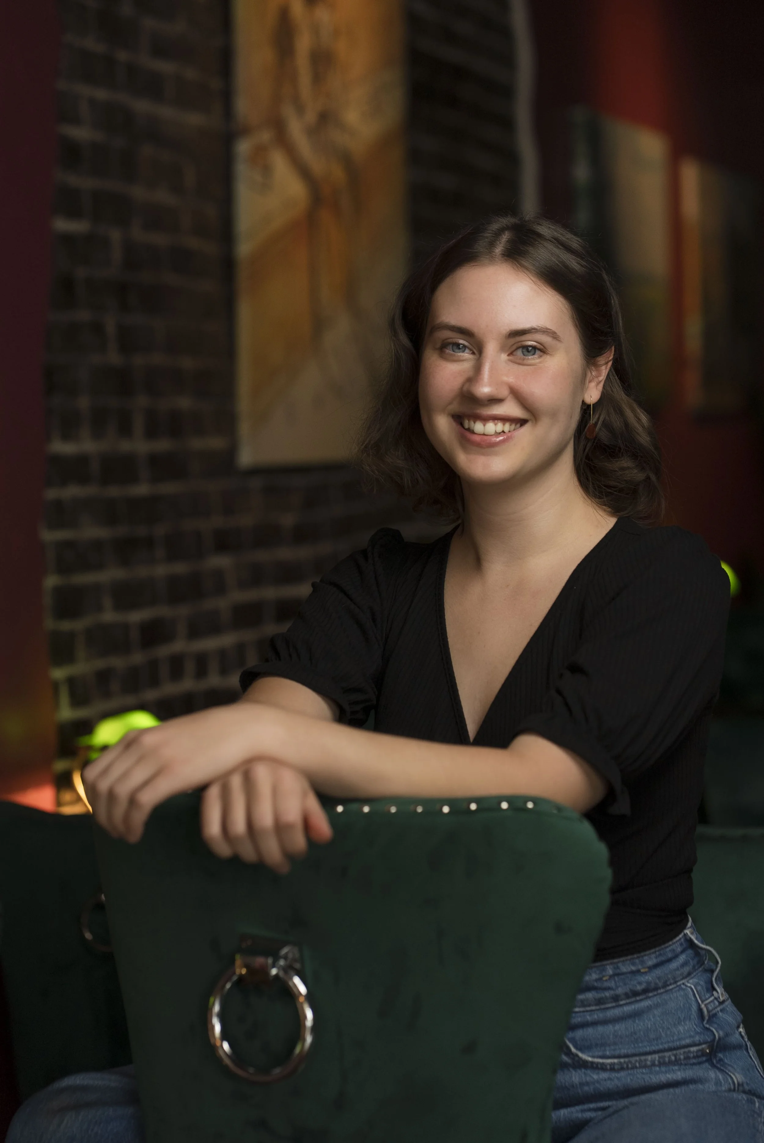 A young woman with shoulder-length brown hair and blue eyes smiling, wearing a black top and earrings, in a dimly lit room with brick wall and artwork in the background.