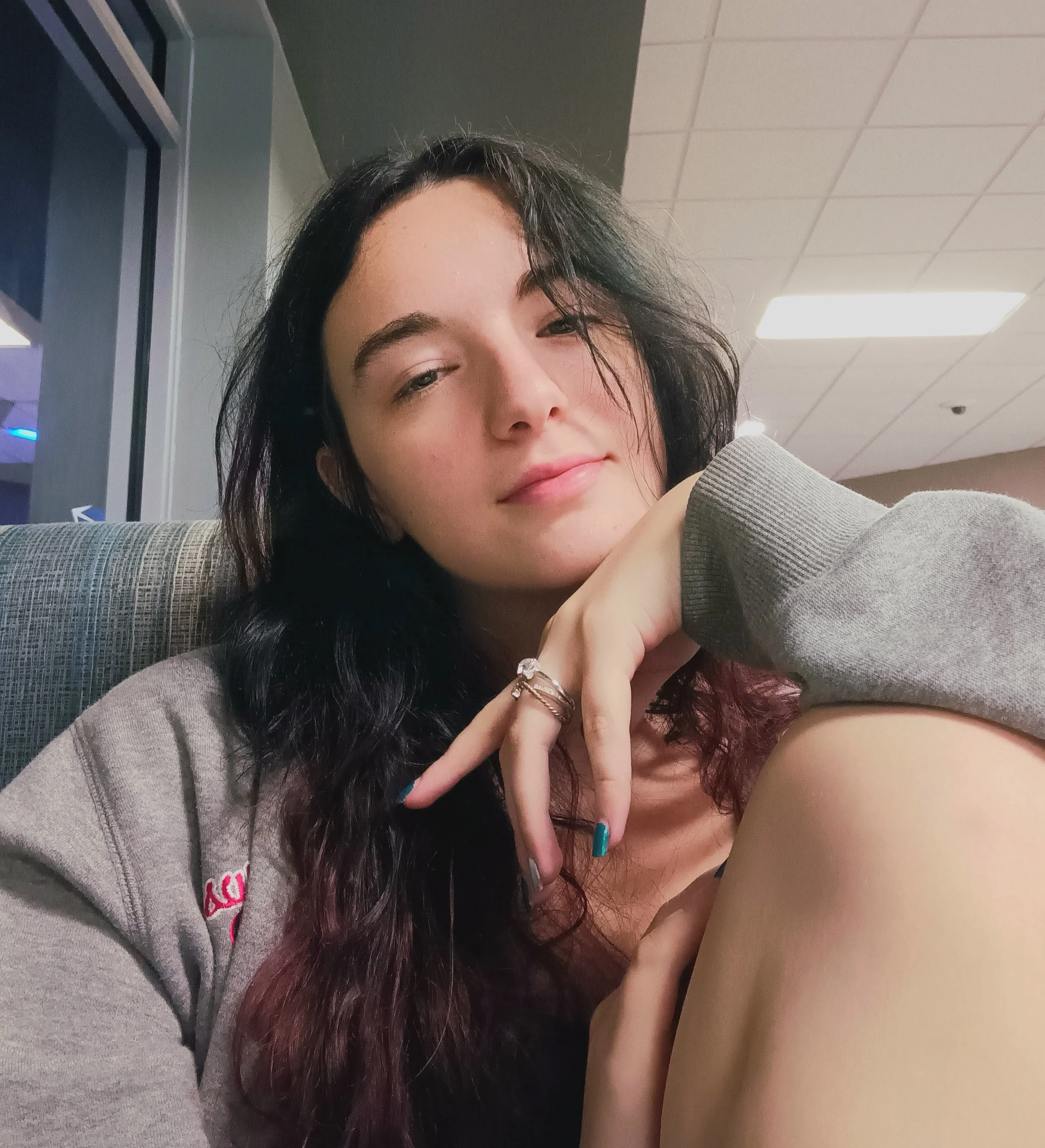 A young woman with dark, wavy hair sitting in a public space, resting her chin on her hand, wearing a gray sweatshirt and a ring, with a neutral expression on her face.