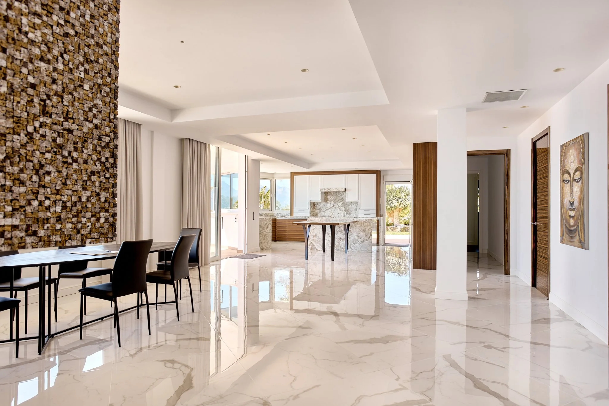 Interiores de una casa moderna con piso de mármol brillante, paredes blancas, y ventanas grandes con cortinas claras. Se observa una mesa de comedor con sillas negras y una zona de cocina con barra de piedra y fondo de vista natural.