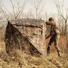 Person setting up a camouflage hunting blind in a field with dry grass and bare trees.