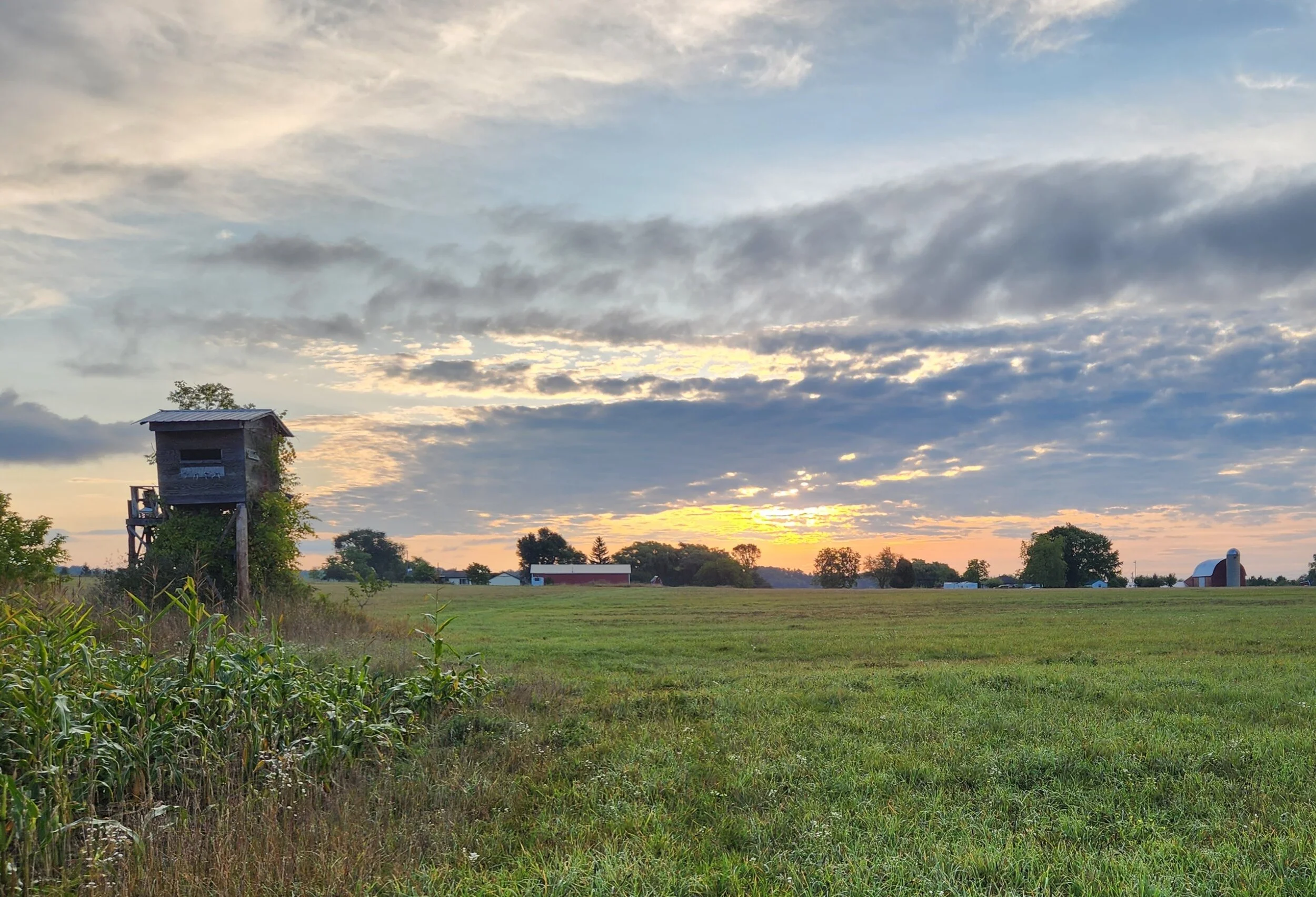 Sunset over a rural landscape with a wooden hunting stand, green field, trees, and a barn in the distance under a cloudy sky.