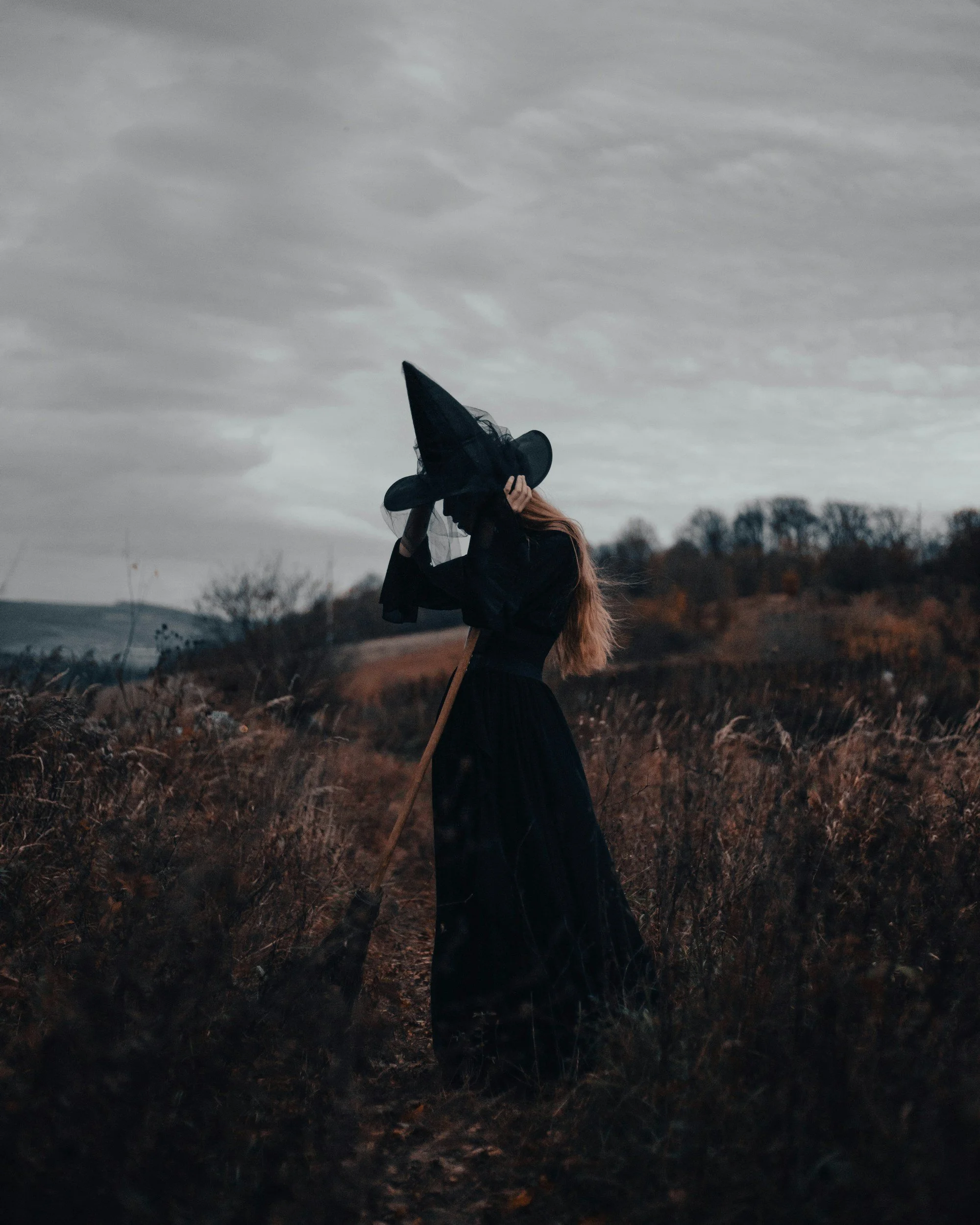 A person dressed as a witch holding a large witch hat, standing outdoors in a field with overcast cloudy sky and trees in the distance.