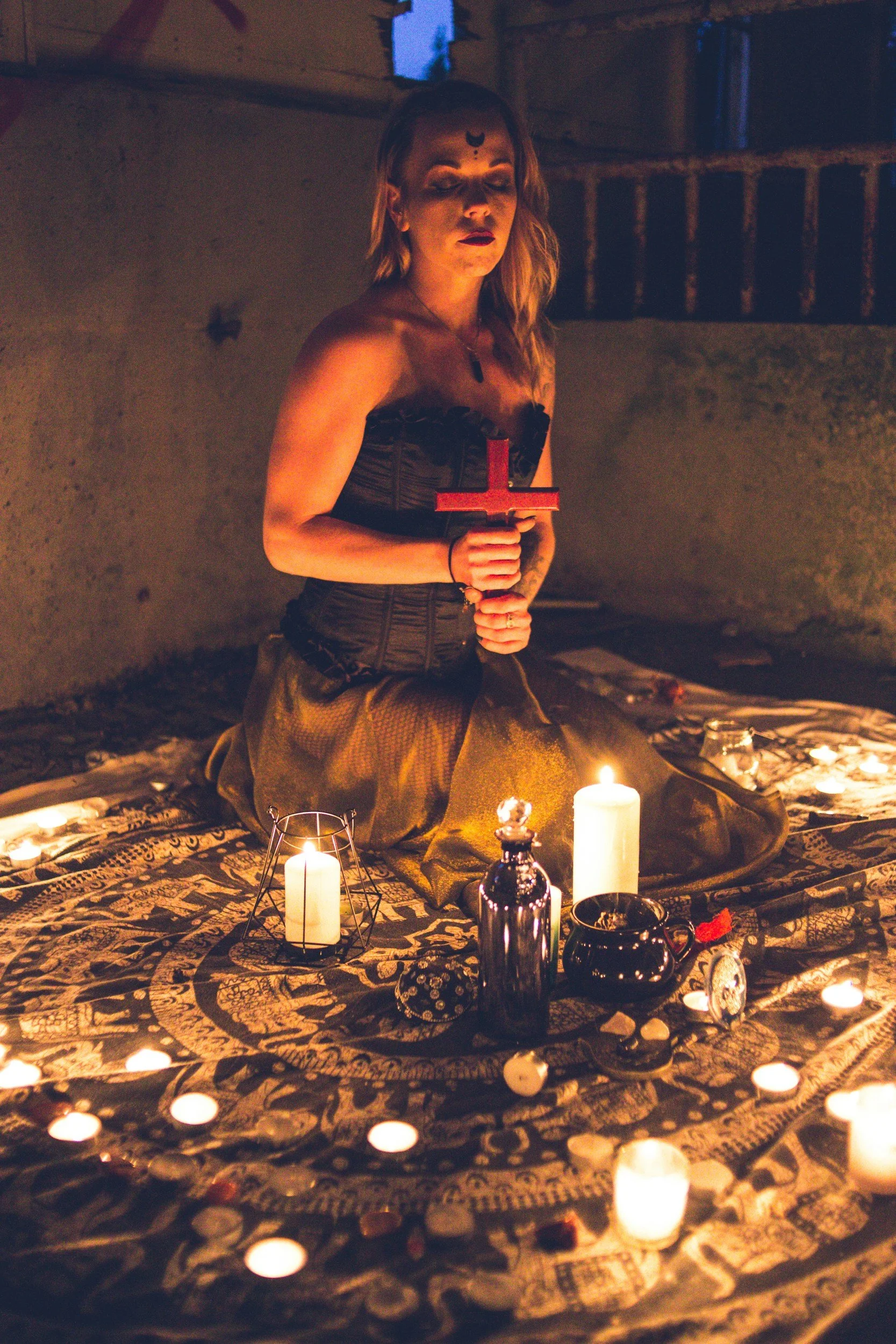 A woman with long, wavy blonde hair, wearing dark makeup and a black strapless top, is kneeling on a decorative cloth with candles and bottles arranged around her. She is holding a red cross in her hands and has a dark symbol on her forehead. The setting appears to be a dimly lit, ritualistic or spiritual space with candles providing warm light.