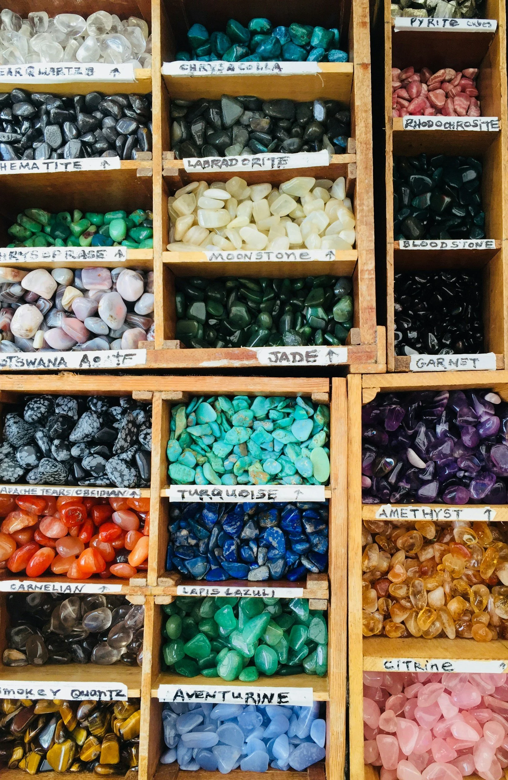 A wooden display box with multiple compartments filled with various colorful gemstones, each labeled with different mineral names such as turquoise, moonstone, amethyst, citrine, and more.