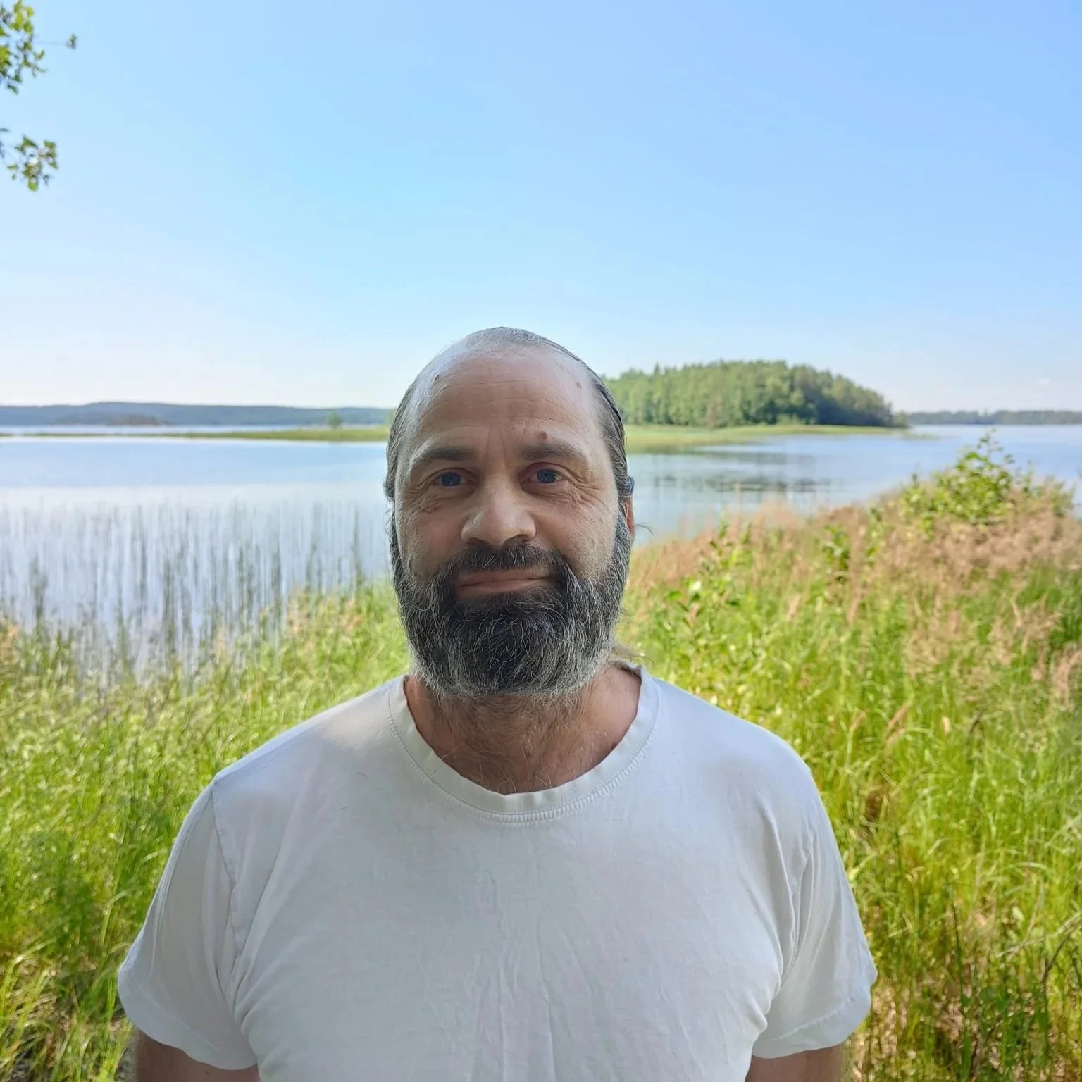 Mies seisoo luonnonkauniilla rannalla järven äärellä, taustalla vesistö ja metsäiset kukkula. Taivas on kirkas ja selkeä.