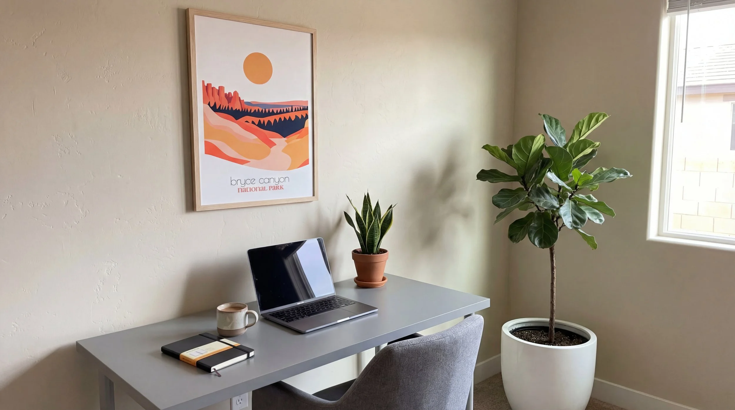 Home office with a gray desk, laptop, notebook, mug, potted snake plant on the desk, large fiddle leaf fig tree in a white planter, and framed abstract art of Bryce Canyon on the wall near a window.