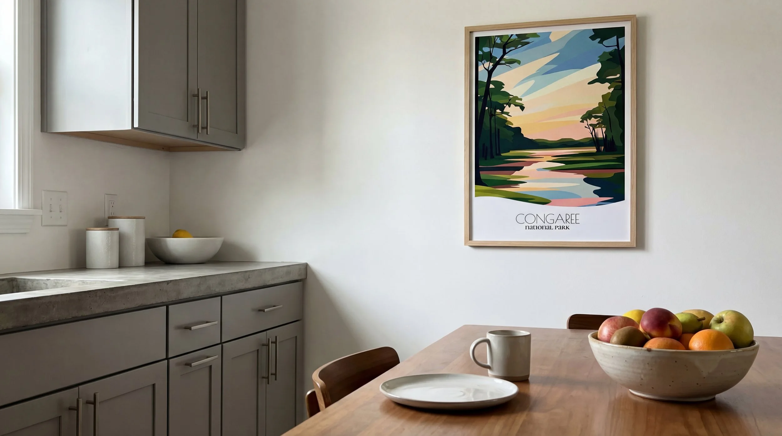 A kitchen with gray cabinets, a concrete countertop, a wooden dining table with a bowl of assorted apples, a mug, and a plate. There is a framed colorful landscape poster of Congaree National Park on the wall.