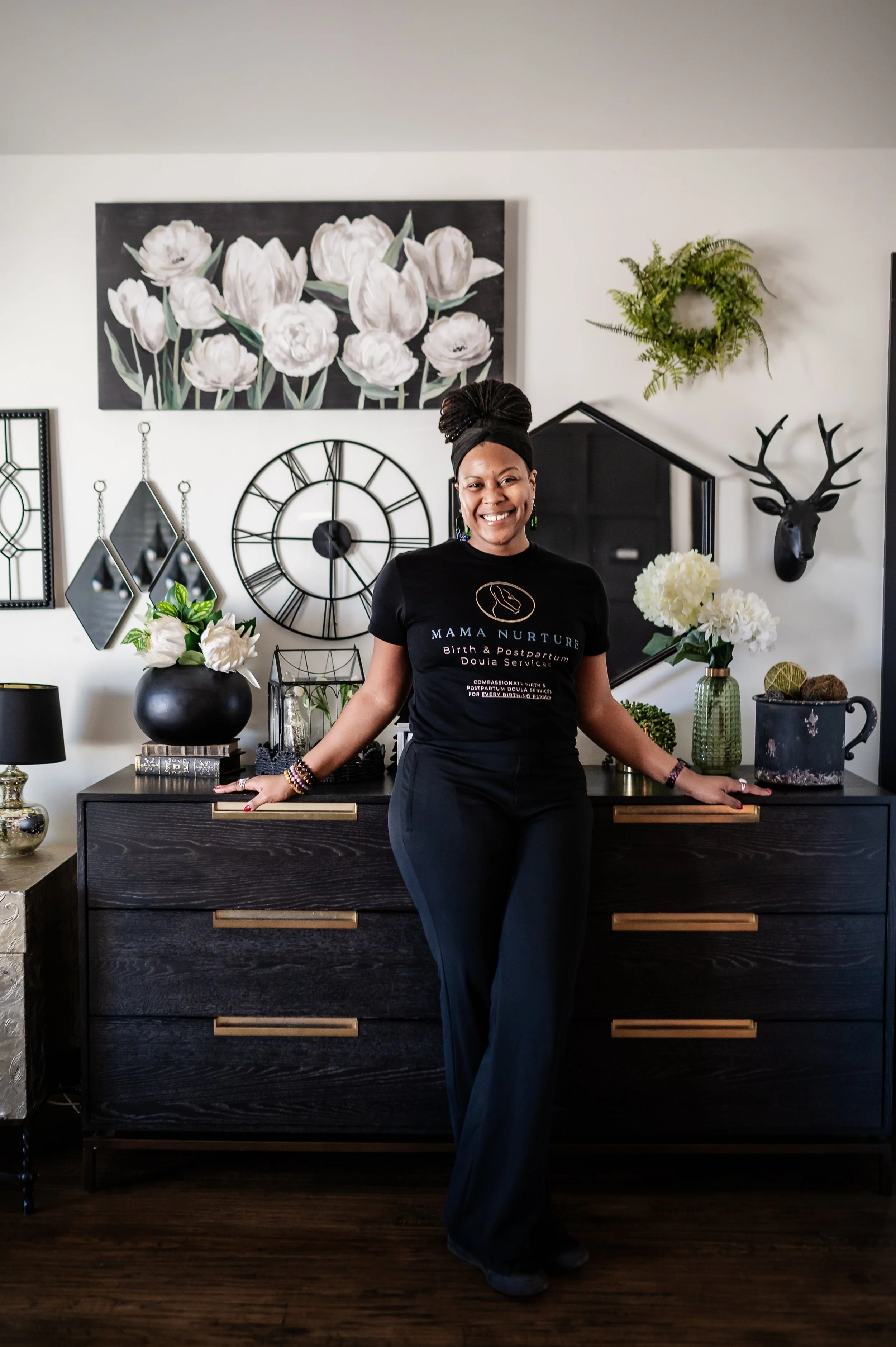 Black doula dressed in a black t-shirt and black pants standing in front of a black dresser with floral decorations behind her.