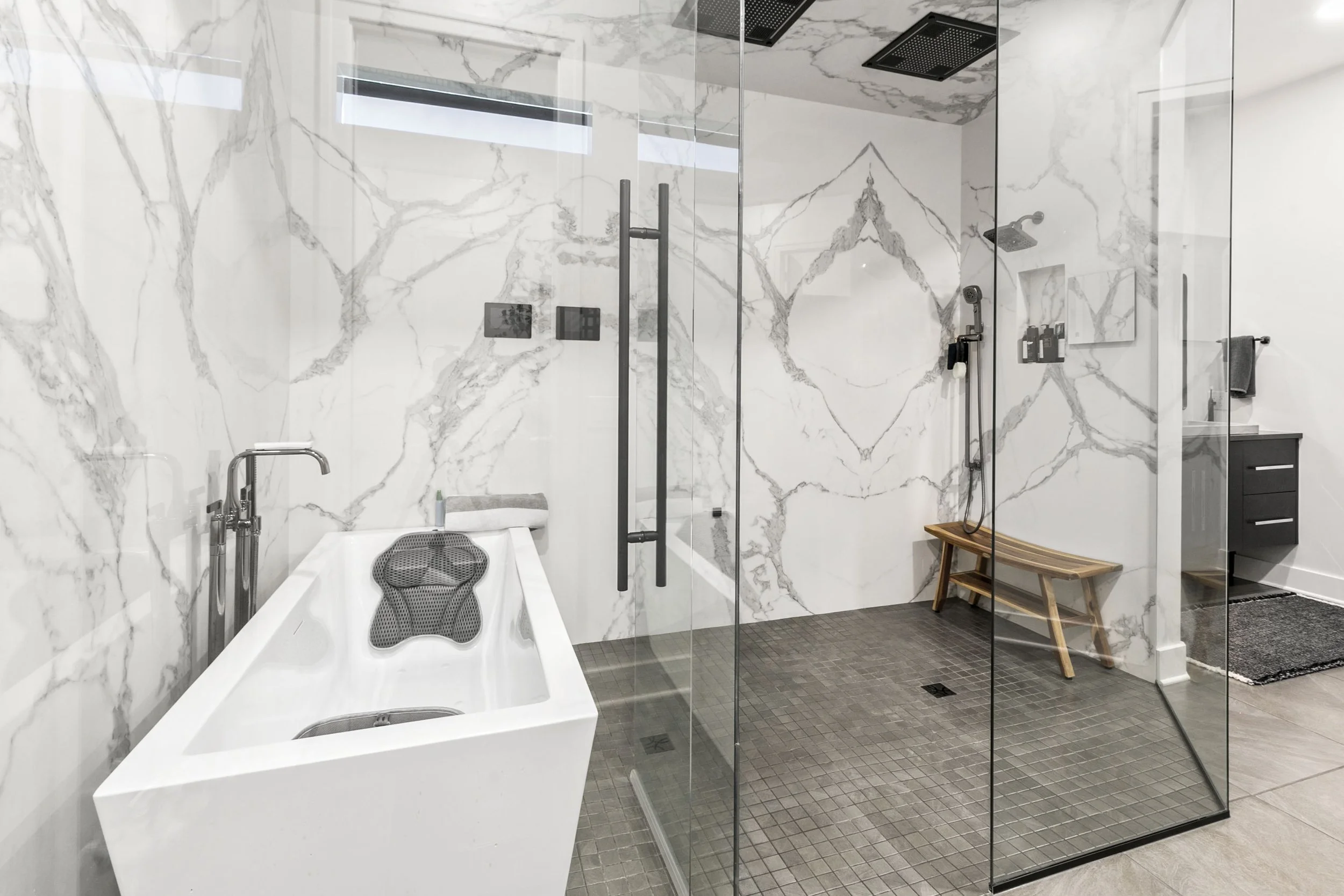 Modern bathroom with a rectangular bathtub, marble wall, glass-enclosed shower area with wooden bench, and a small dark vanity with a towel