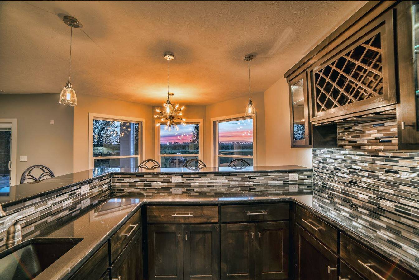 View of a modern kitchen with dark cabinets, a tiled backsplash, pendant lights, and a sunset visible through large windows.