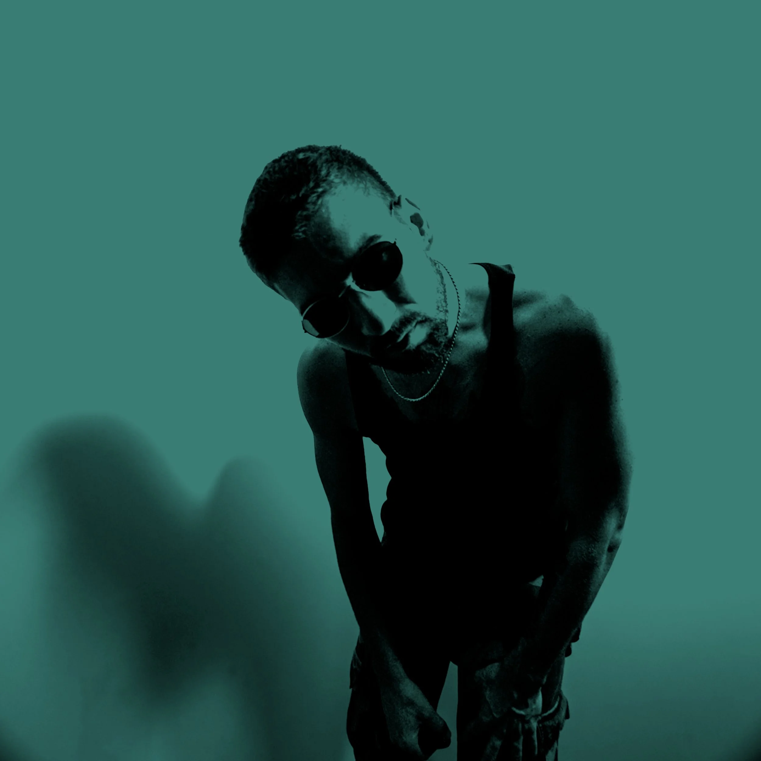 A man wearing sunglasses, a black tank top, and a chain necklace leaning forward with a shadow cast on a plain wall.