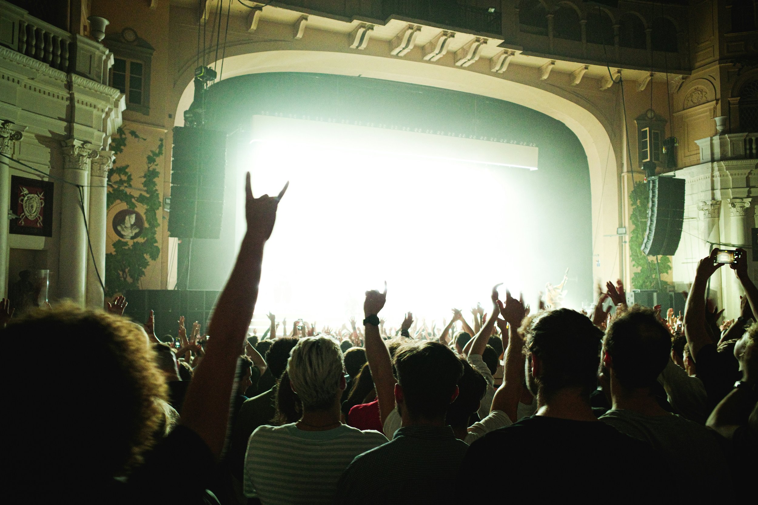 Concert crowd with raised hands and one showing rock hand gesture, in front of brightly lit stage inside an ornate theater.
