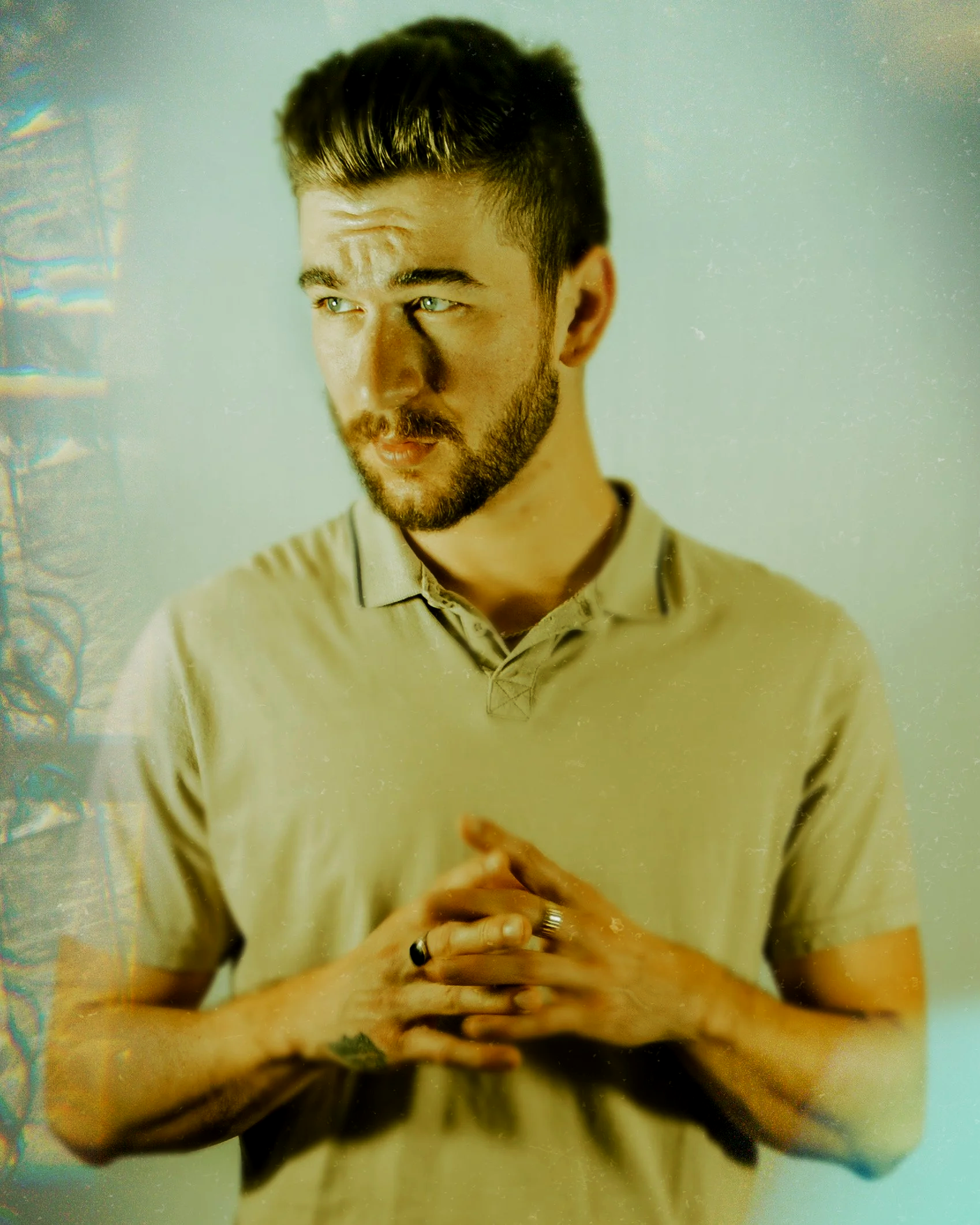 A young man with short dark hair and a beard, wearing a beige polo shirt, standing with his hands clasped together in front of him, looking thoughtfully to his left against a plain, light-colored background.