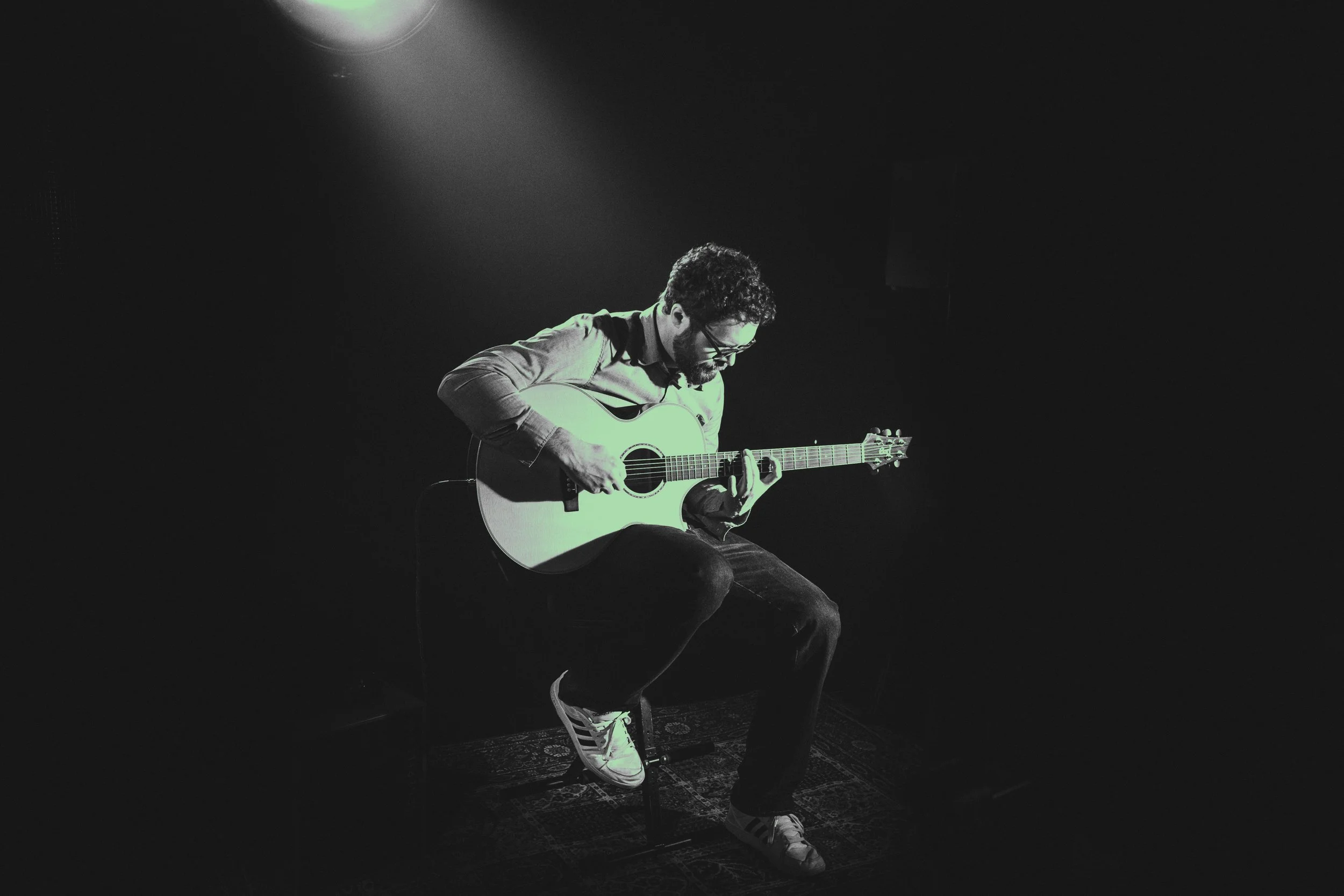 A man seated on a stool playing an acoustic guitar on a dark stage, illuminated by a single spotlight.