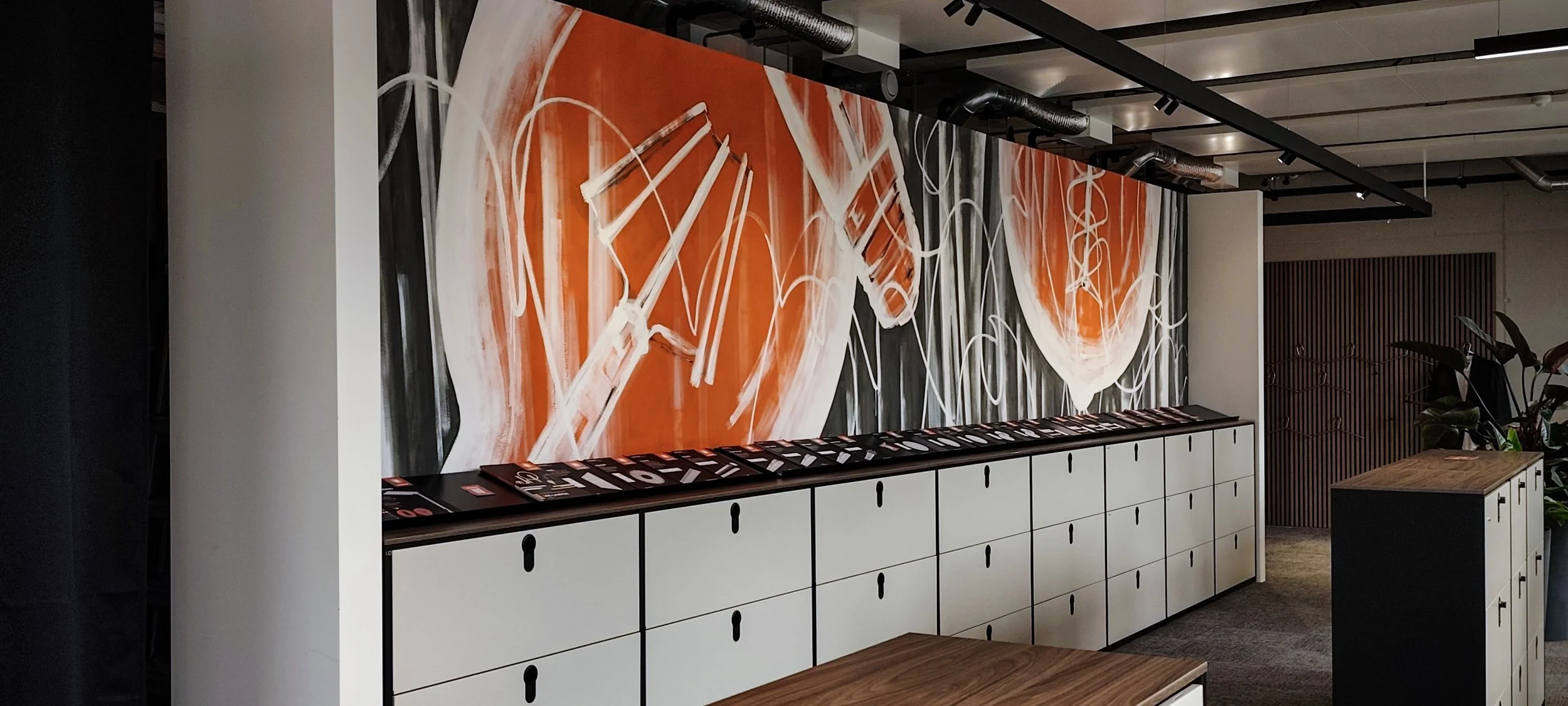 Modern interior with a large abstract wall art featuring orange, black, white, and gray colors, above a white storage cabinet with black handles and magazines on top. There is a small wooden table in the foreground and some plants to the right.