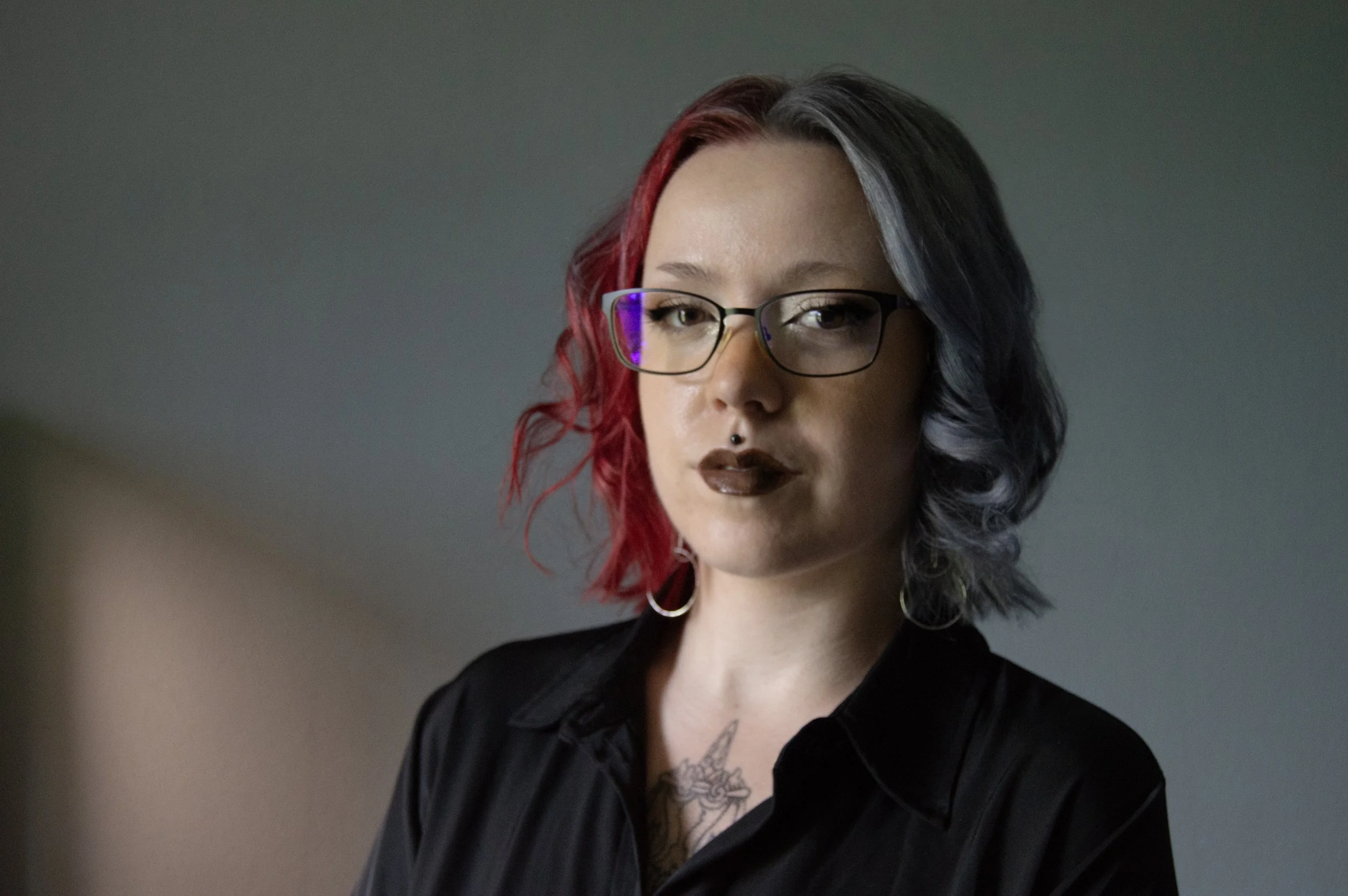 A young woman with shoulder-length hair, half dyed red and half dyed black, wearing glasses with purple reflections, dark lipstick, earrings, and a black shirt, looking directly at the camera against a plain gray background.
