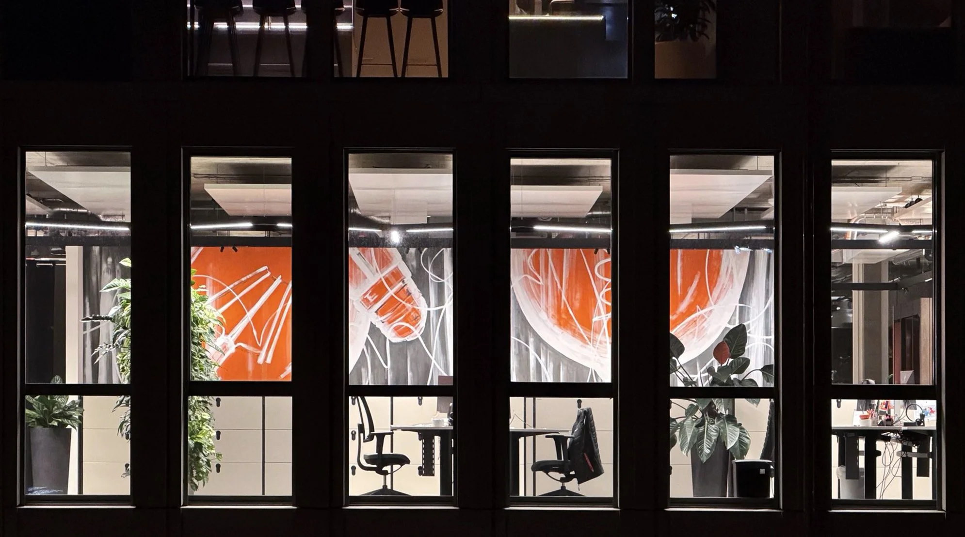 View through large window showing an interior office space with chairs, plants, and a bright orange abstract mural on the wall.