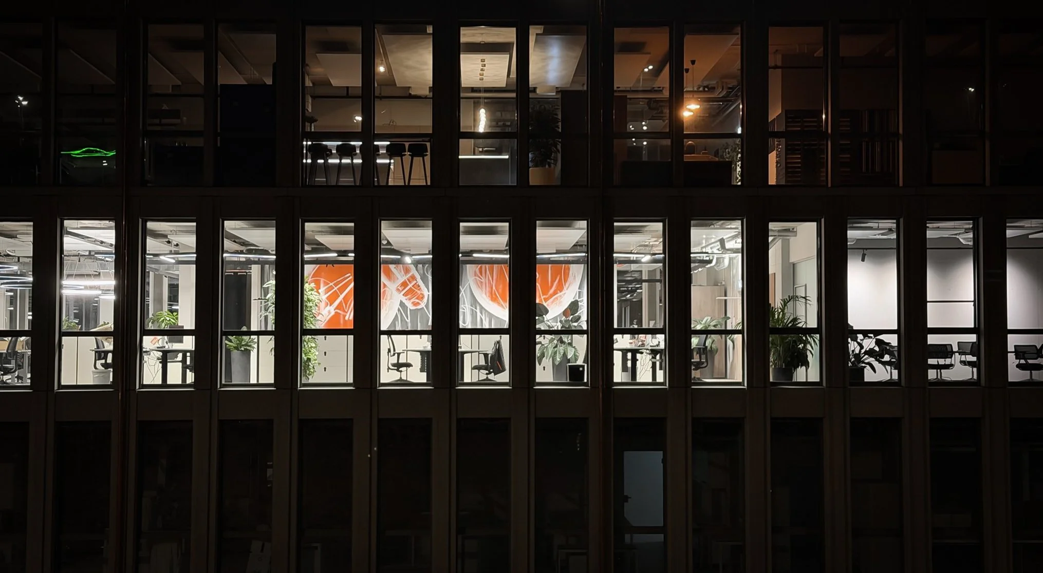 Night view of an office building with illuminated interior, seen through large glass windows, showing workspaces, plants, and abstract wall art.